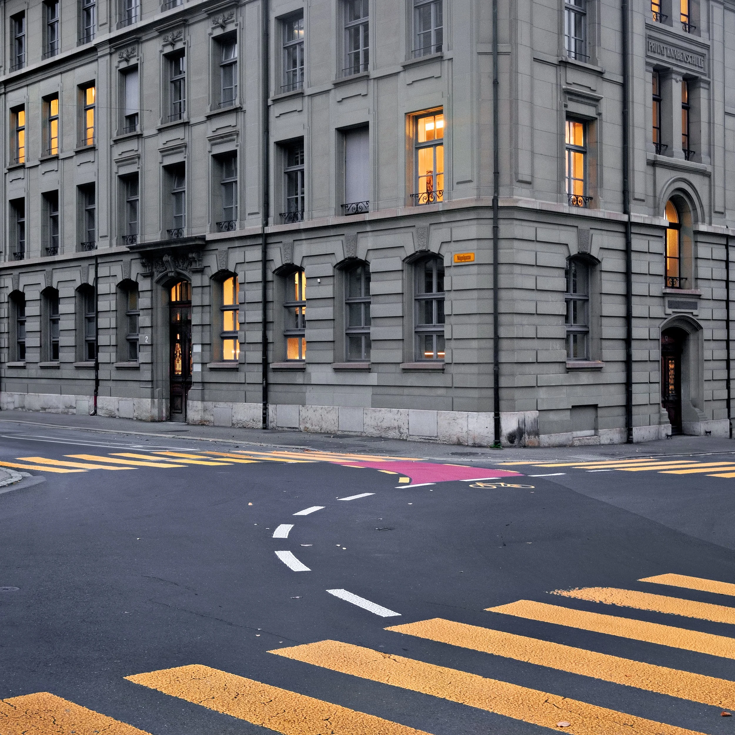 City street corner with a gray multi-story building, illuminated windows, and yellow and pink pedestrian crosswalk markings.
