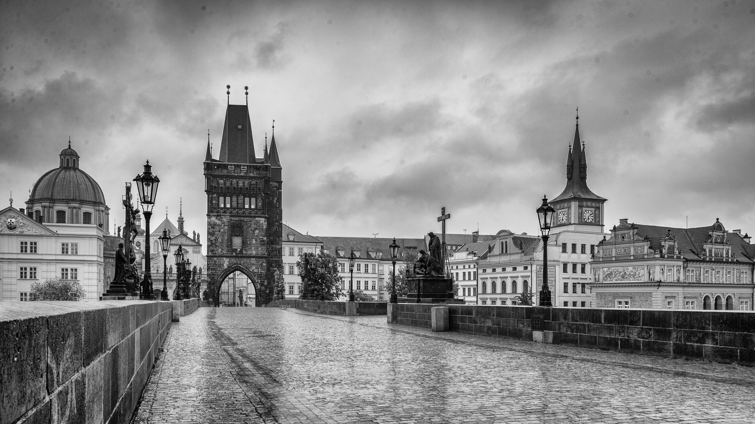 A historic European cityscape in black and white, with a cobblestone bridge, several statues, ornate buildings, church steeples, and a clock tower under a cloudy sky.
