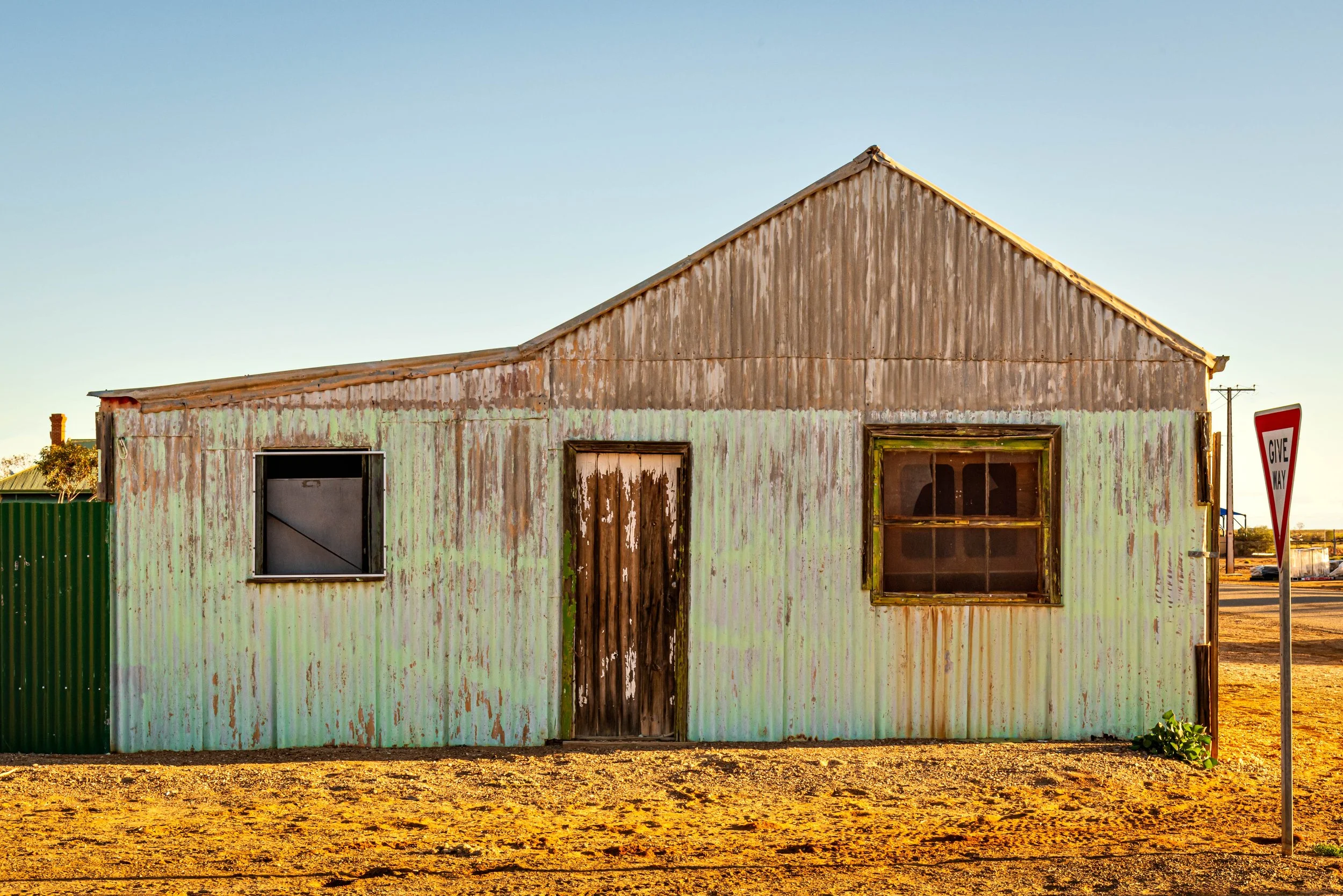 Old weathered house with peeling green paint, metal siding, two windows, and a central door, near a 'Give Way' traffic sign on a dirt lot.