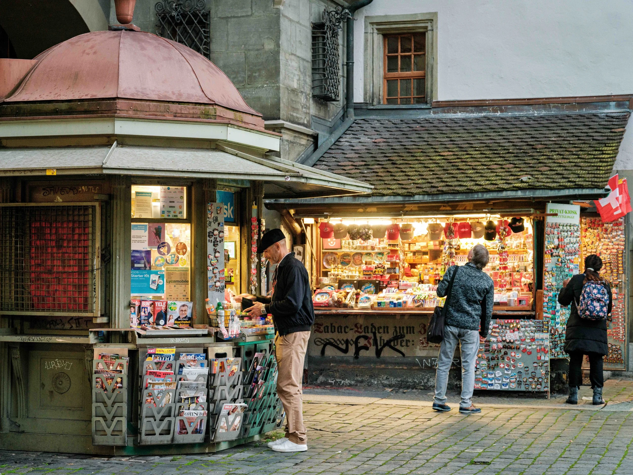 Street newsstand and kiosk with three people browsing, colorful items like badges and souvenirs, brick pavement, old building facade, and Swiss flag.
