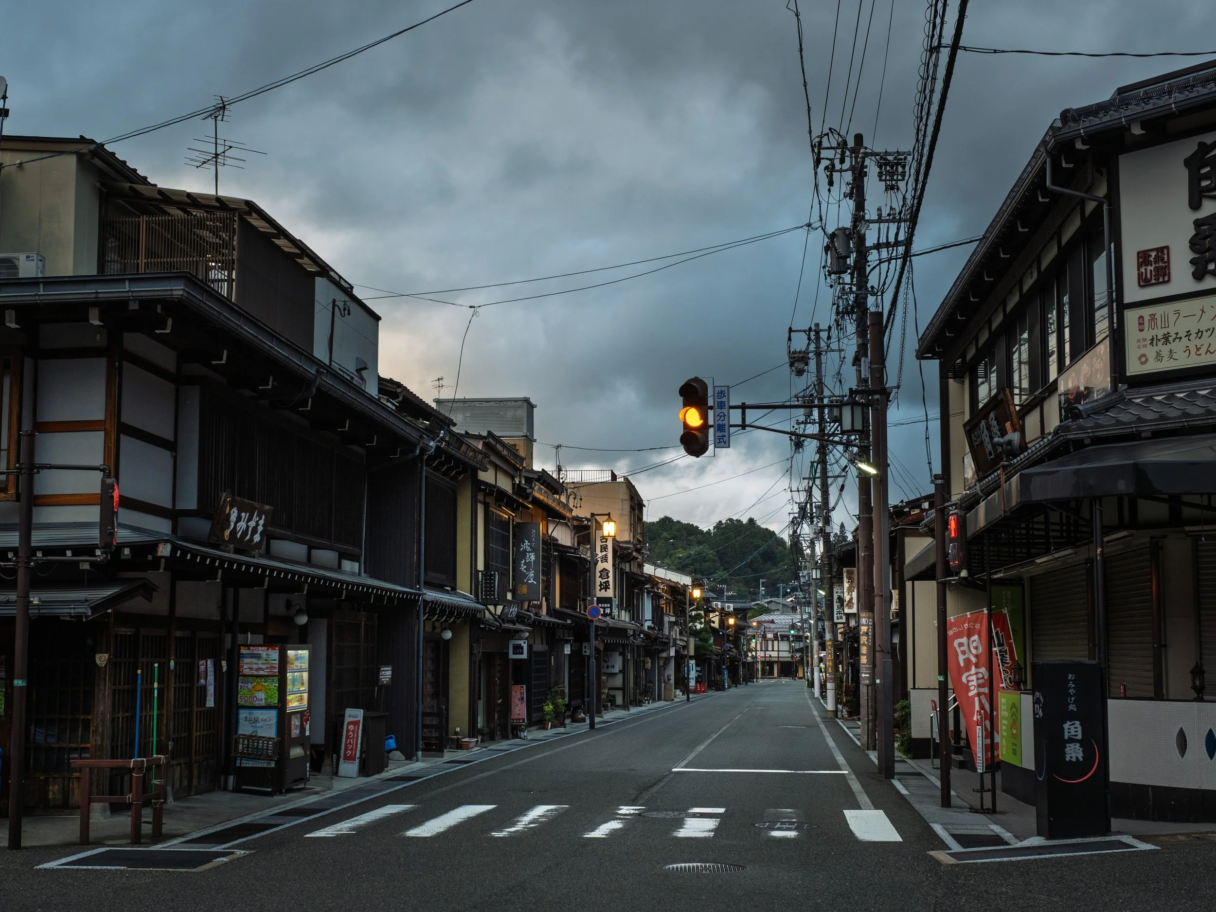 Empty street in a Japanese town with traditional wooden buildings, power lines, traffic lights, and cloudy sky.