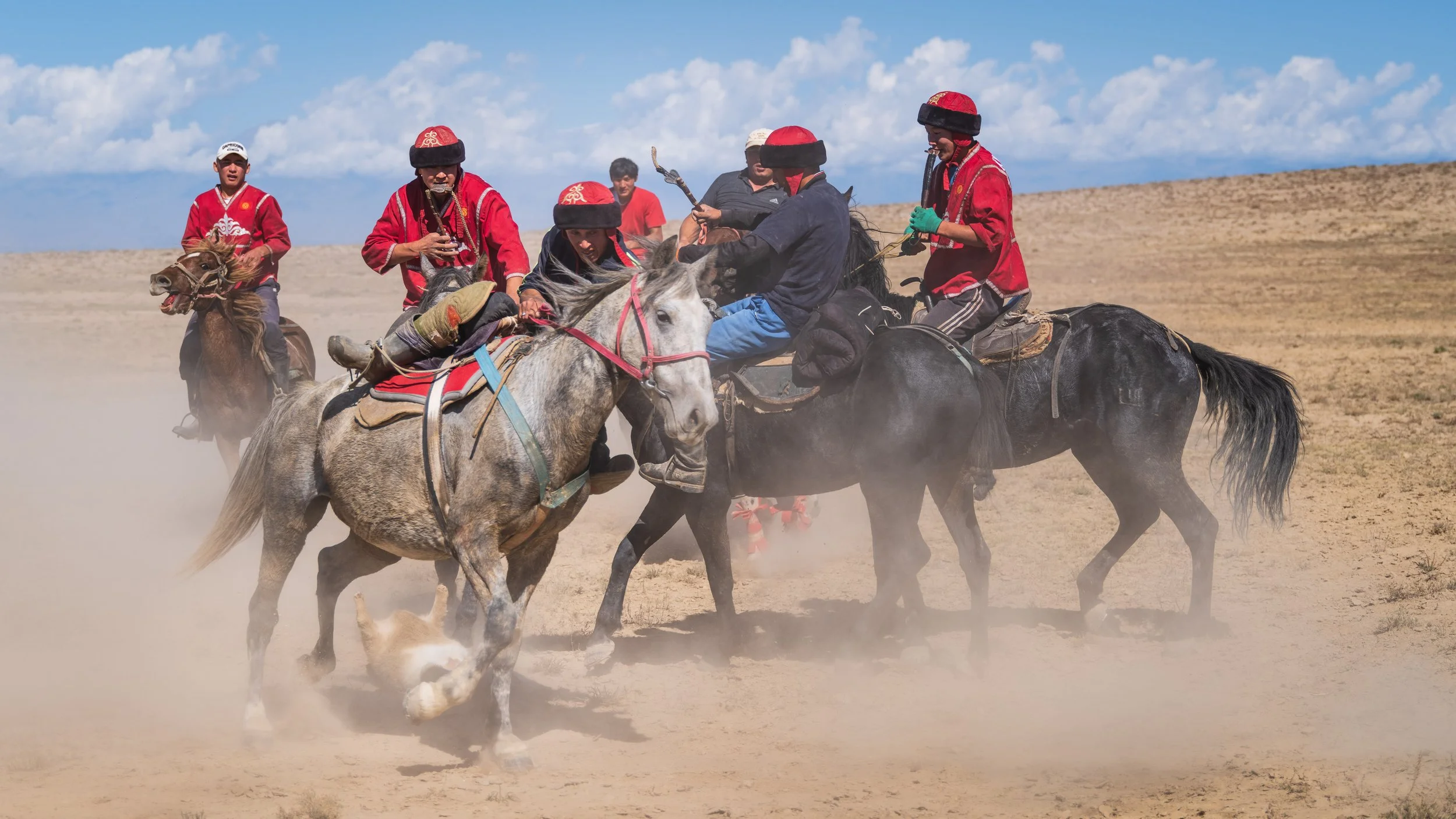 Group of people riding horses and playing a traditional sport on a dusty plain under a blue sky with clouds