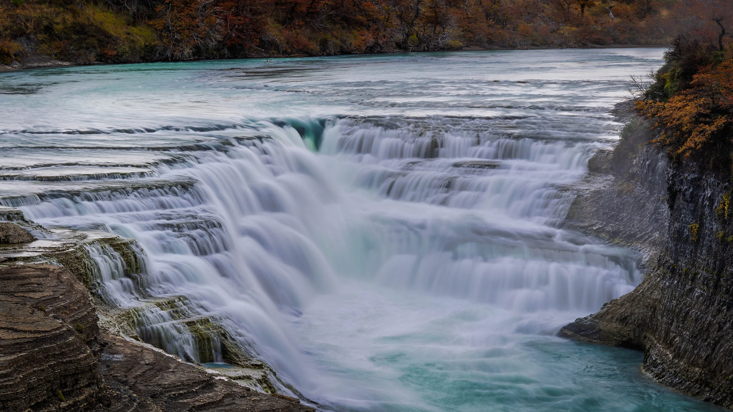 A waterfall cascading over rocks into a river surrounded by trees with autumn foliage.