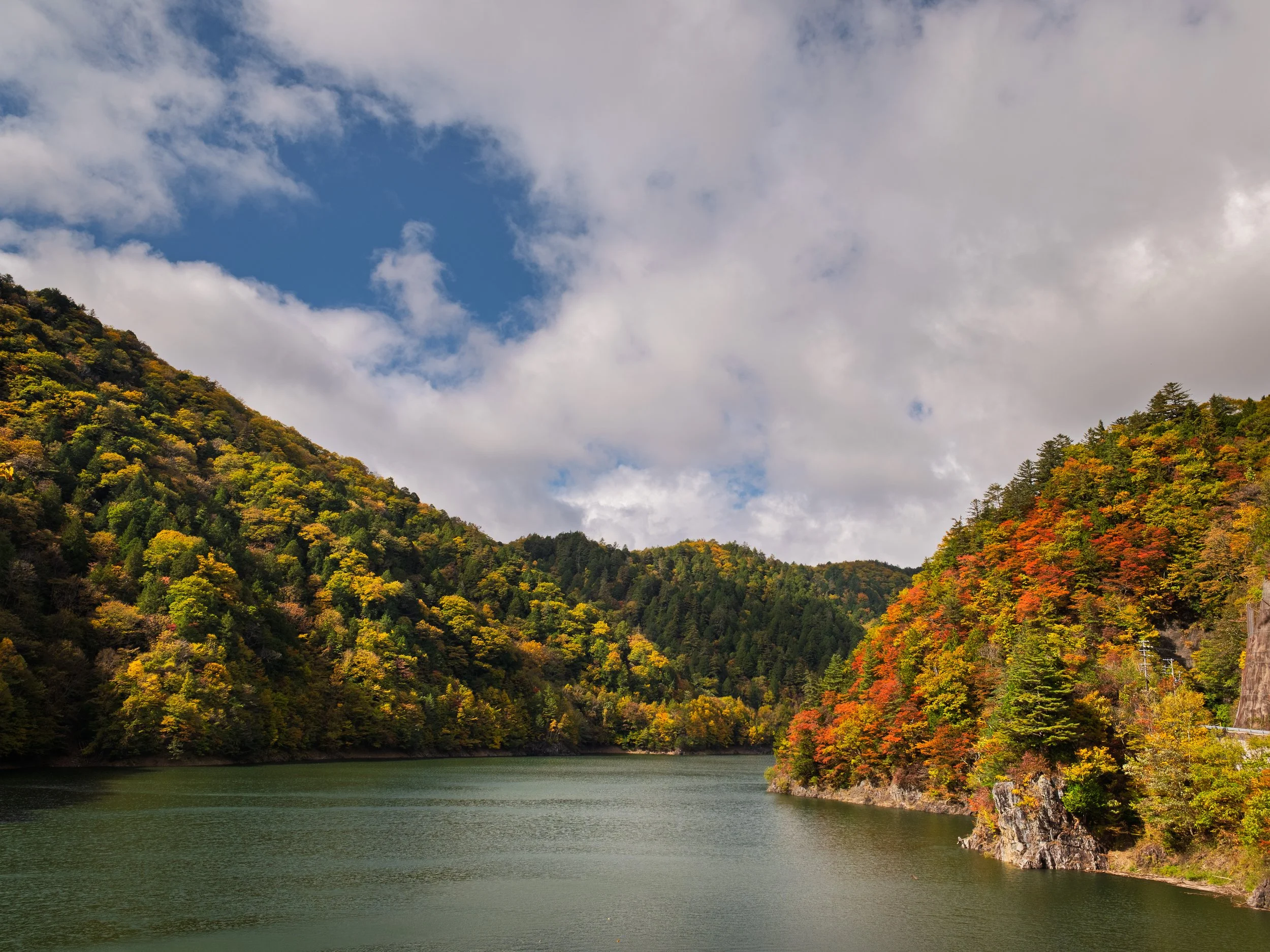 A scenic view of a river surrounded by forested hills with trees showing fall colors under a partly cloudy sky.