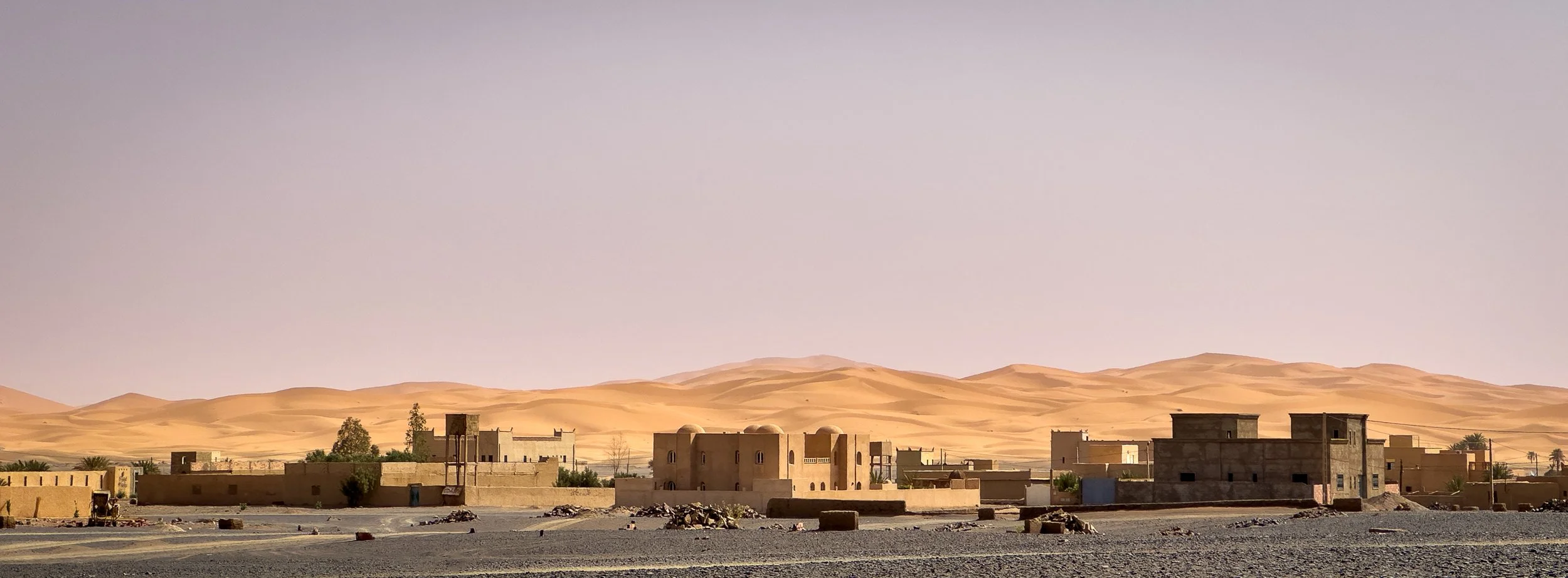 A desert village with sand dunes in the background, featuring rectangular buildings made of mud or clay, some with domed roofs, under a clear sky.