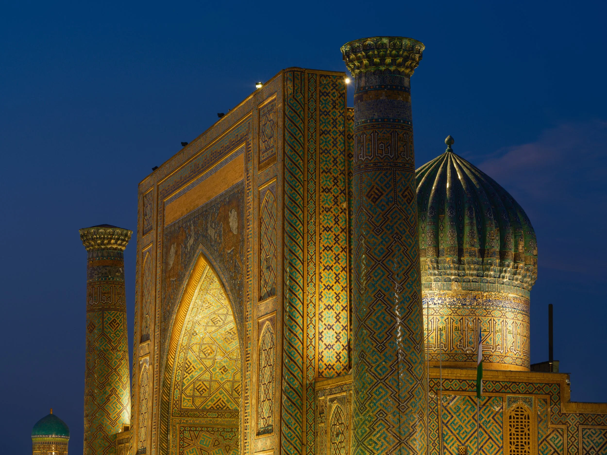 Night view of an elaborately decorated historic building with Islamic architecture, featuring colorful tile work, a domed roof, and tall columns, illuminated against the dark sky.