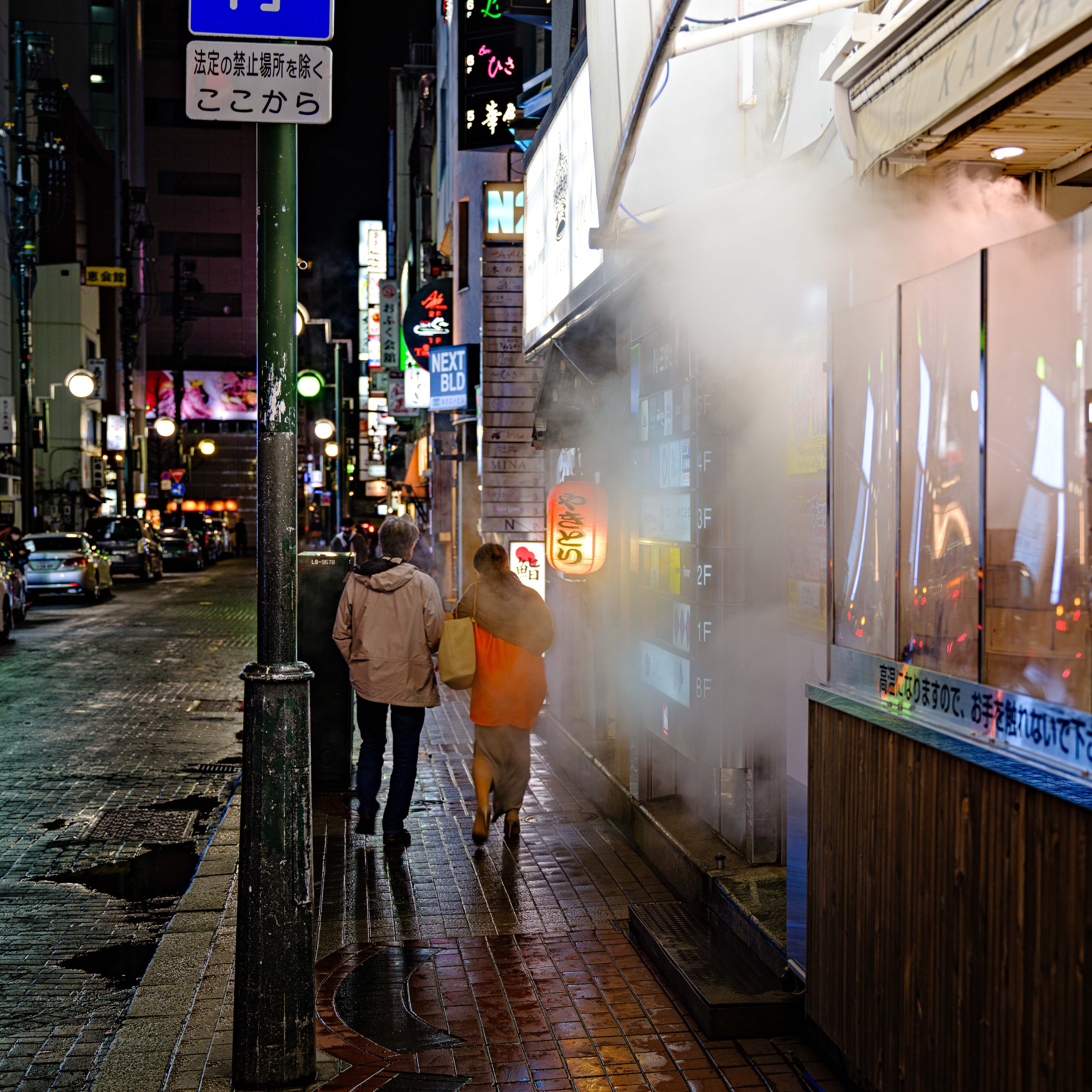 A city street at night with two pedestrians walking past a steamed-up storefront decorated with neon signs and colorful lights.