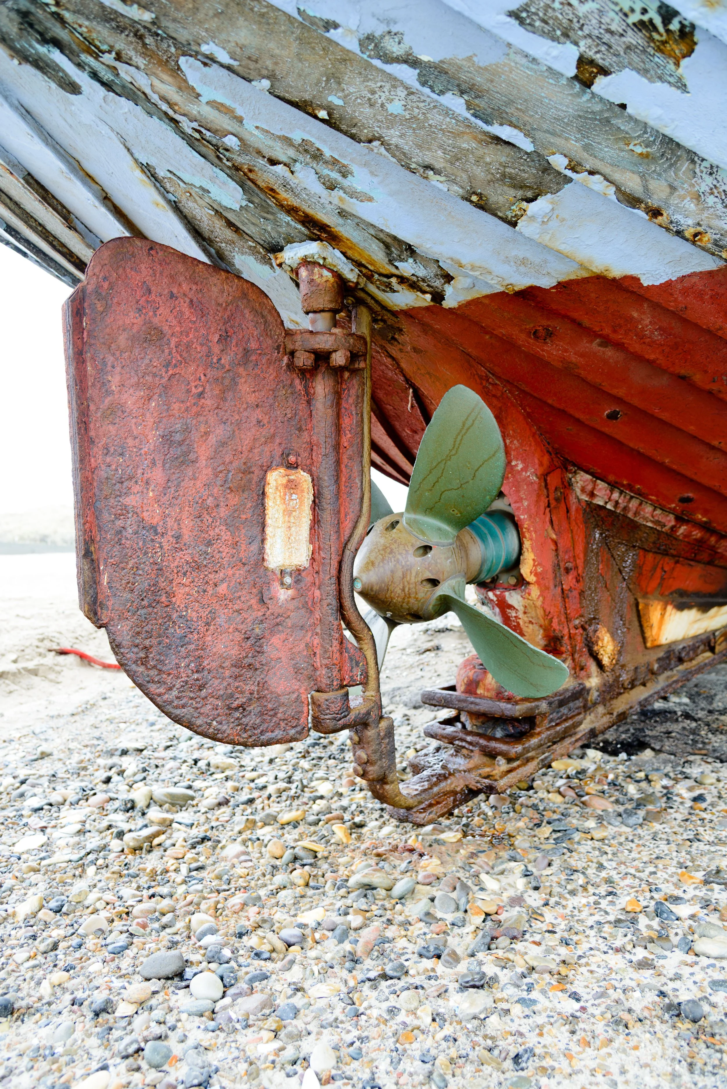 Close-up of a rusty boat hull with a weathered propeller and rudder, resting on a rocky beach.