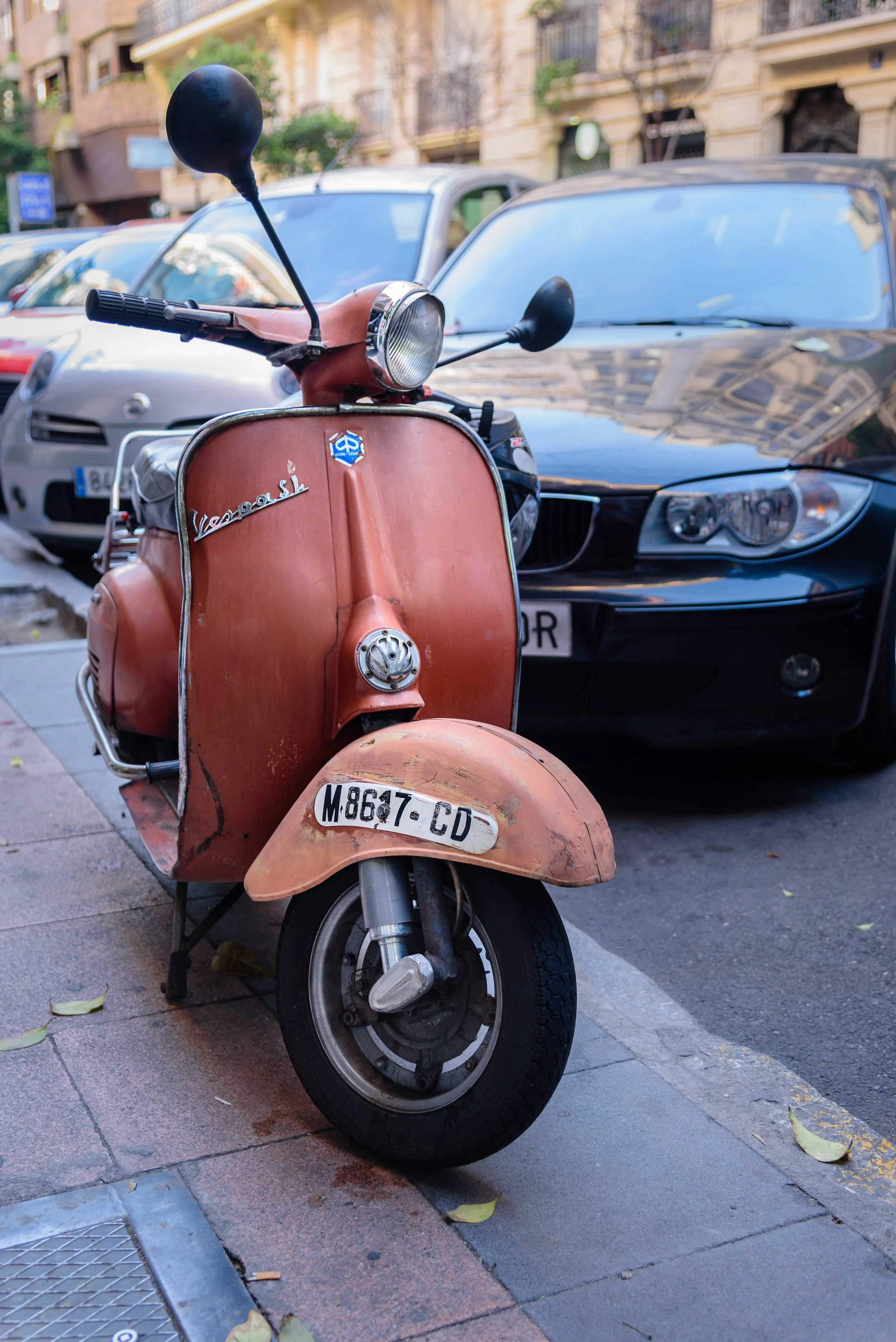 An old rusty orange Vespa scooter parked on the city sidewalk next to a black car and other vehicles, with buildings in the background.
