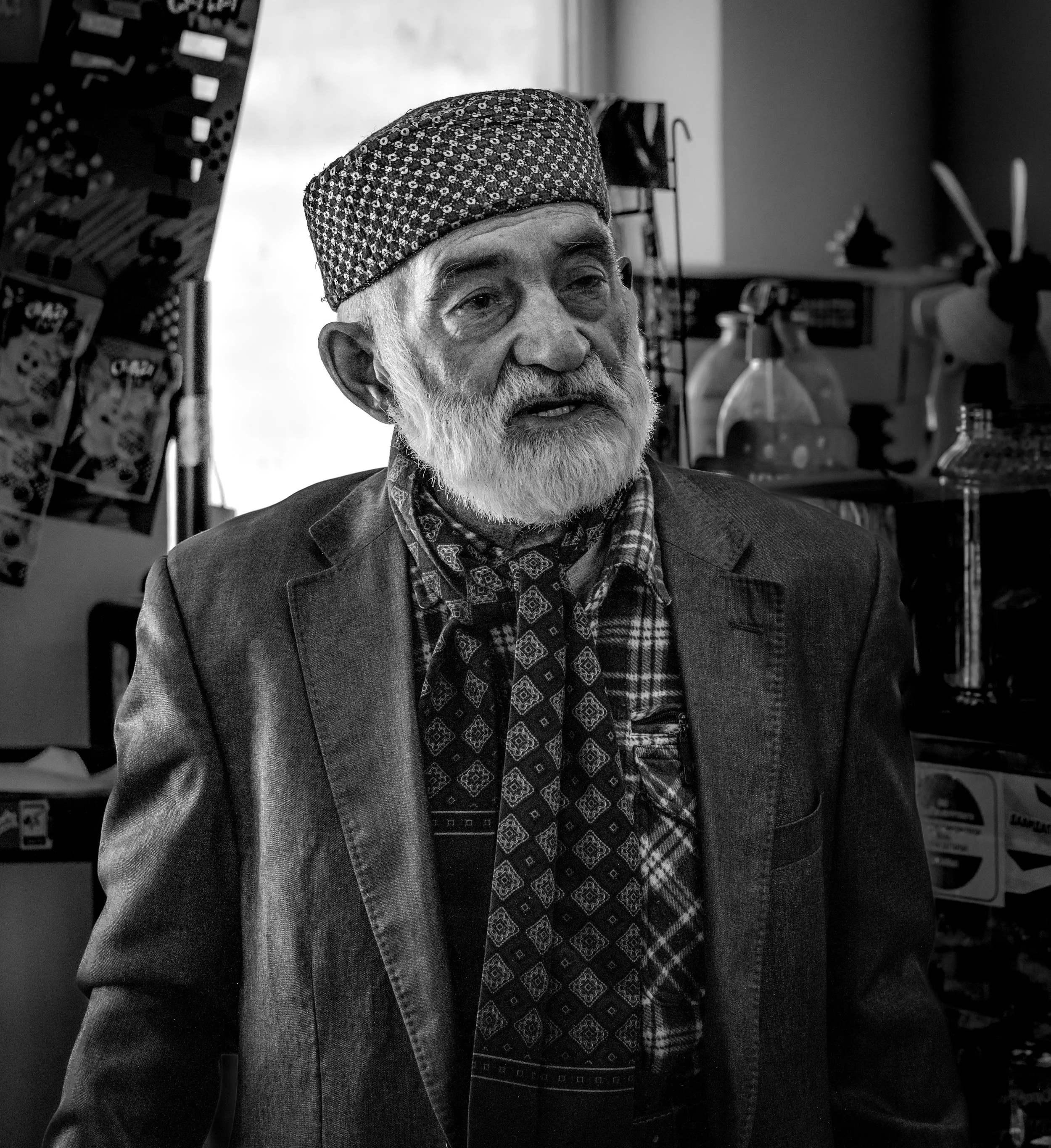 Black and white photo of an elderly man with a beard wearing a patterned cap, plaid shirt, patterned tie, and blazer, inside a store with assorted items on shelves and walls.