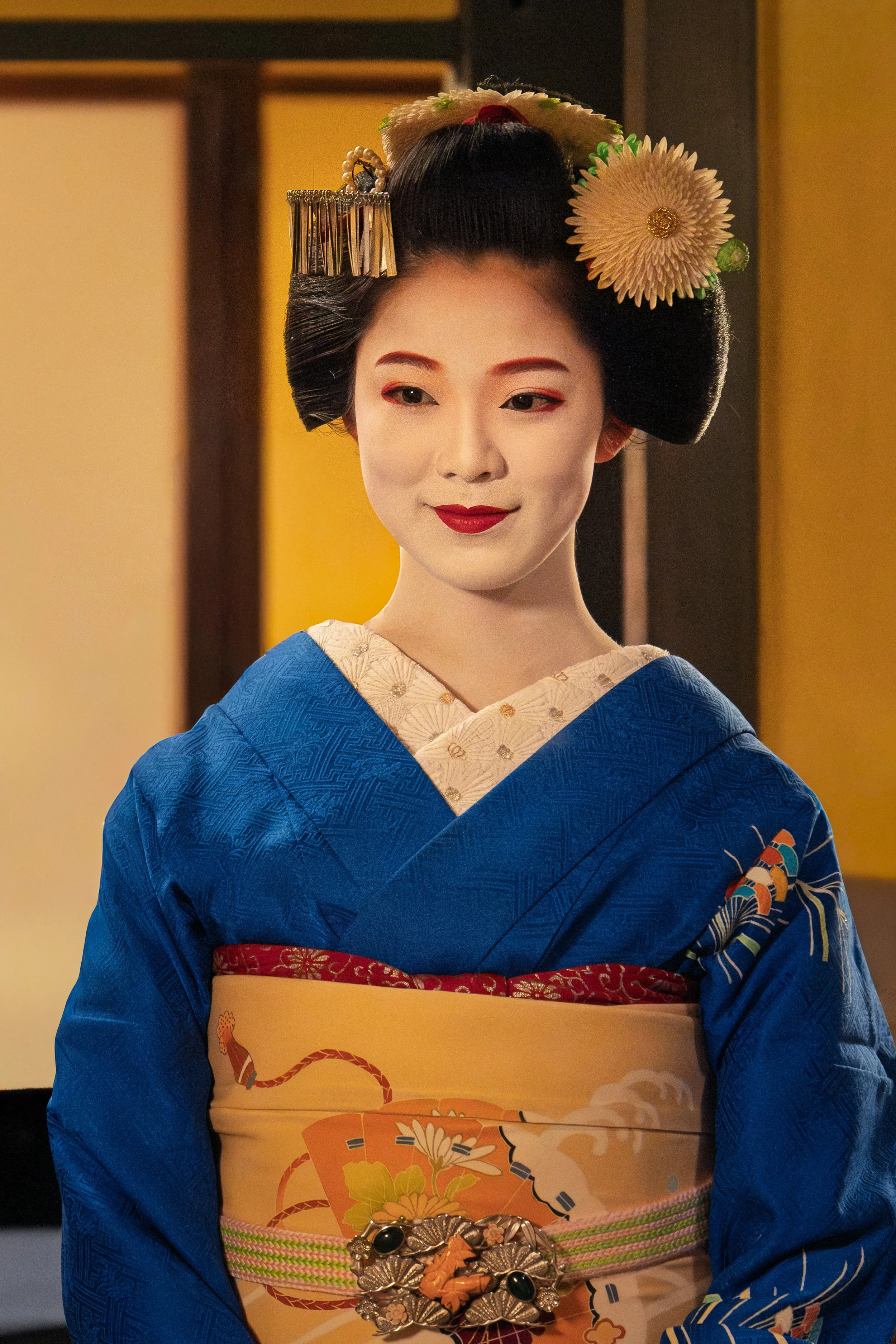 A woman dressed as a traditional Japanese Geisha wearing a blue kimono with intricate patterns and a yellow obi with floral designs, adorned with a large floral hairpiece.