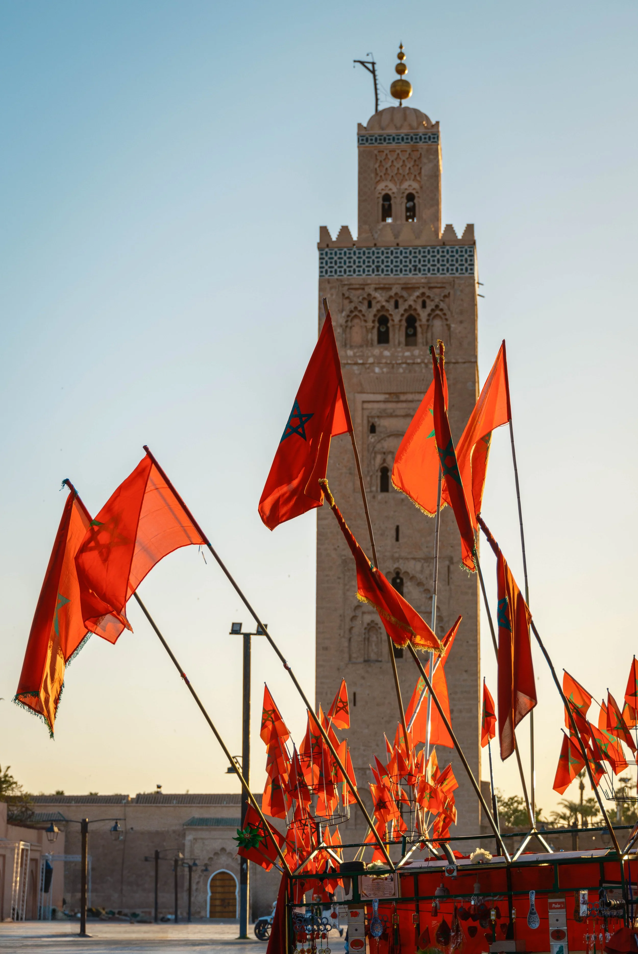Several Moroccan flags with red backgrounds and green stars, waving in front of a tall minaret during daylight, with the sky clear and blue.