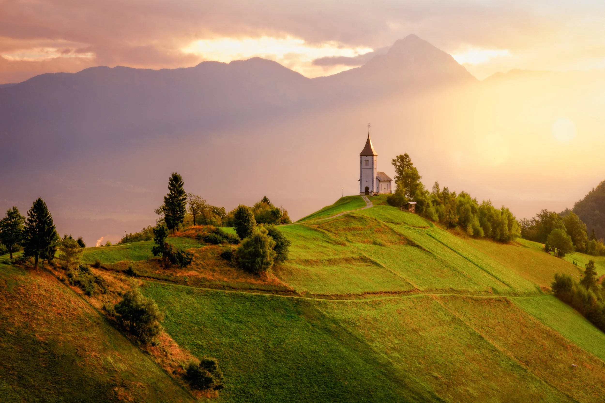 A small white church with a tall steeple on a grassy hilltop during sunset with mountains in the background.