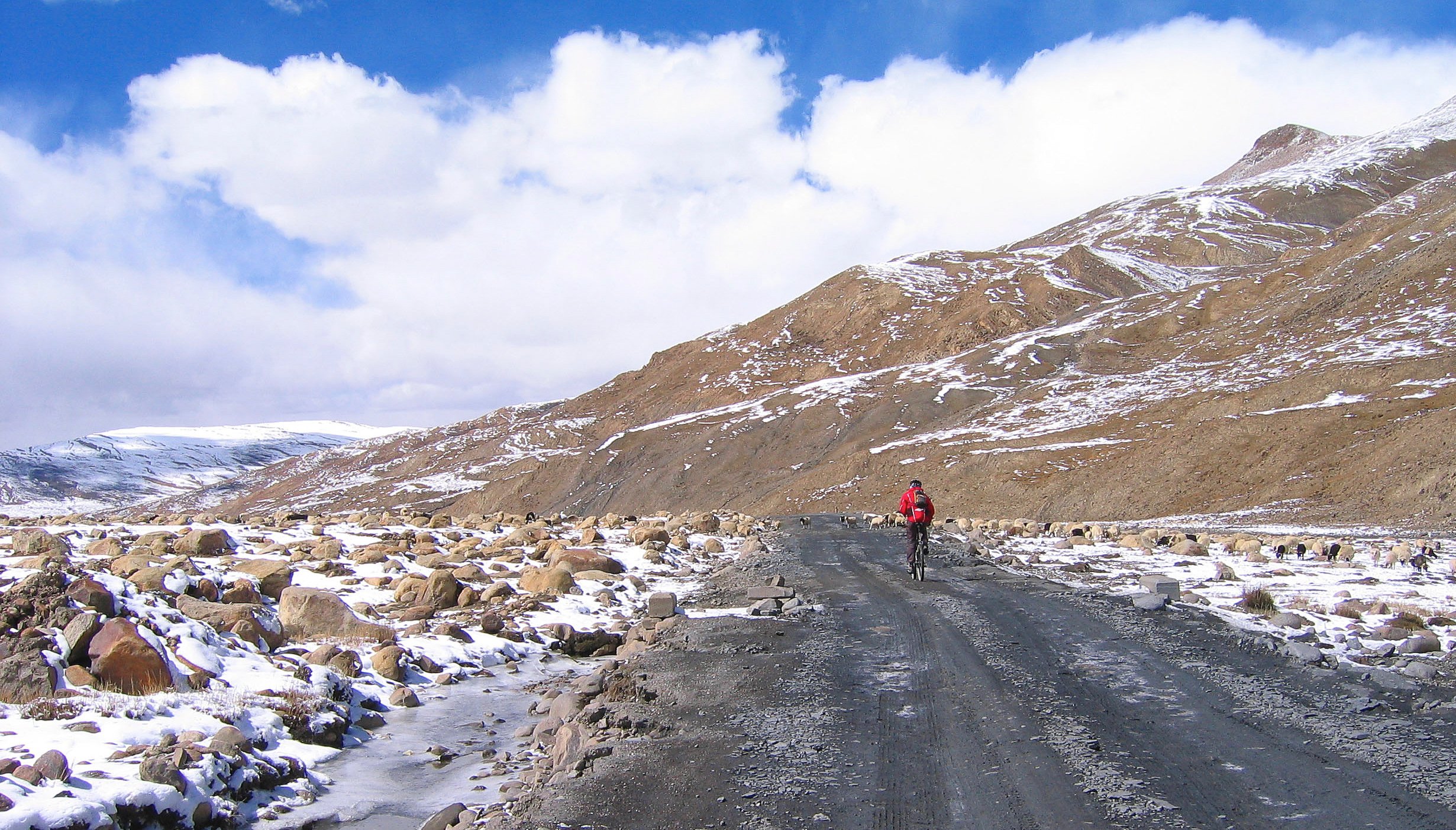 A person in a red jacket biking on a rocky, muddy trail through a snowy mountainous landscape with scattered rocks and a partially frozen stream.