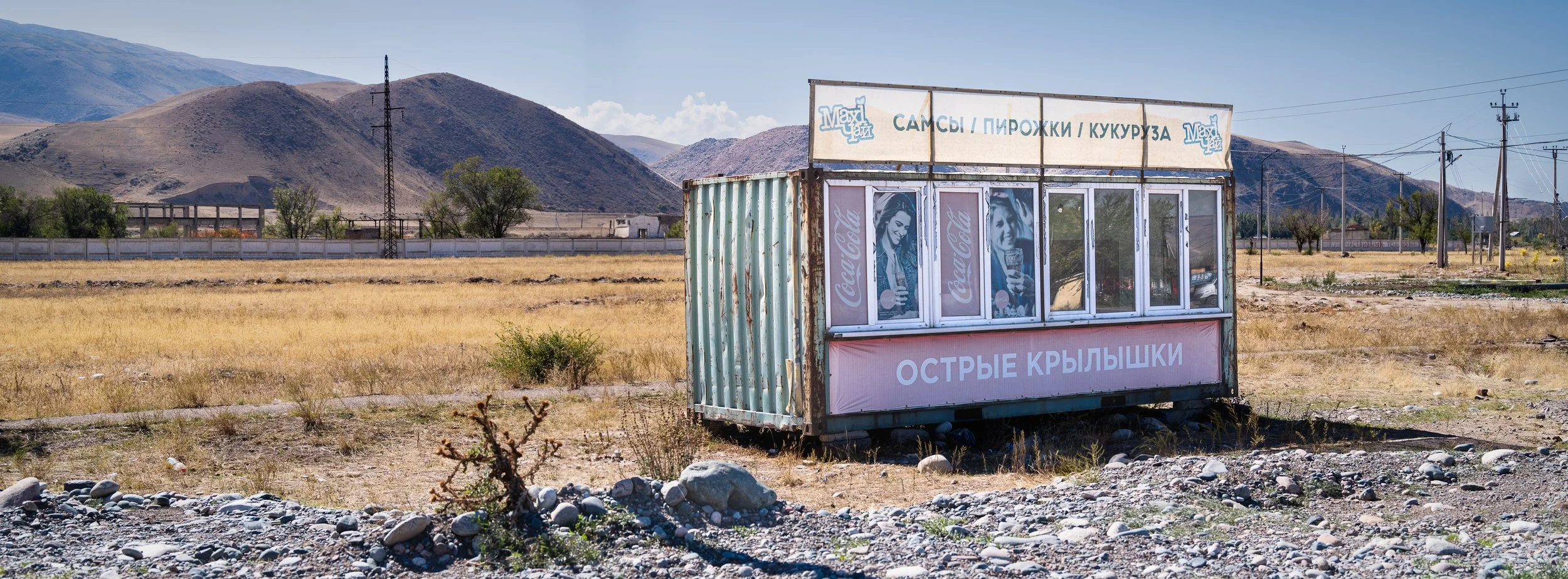 A small, weathered kiosk with Coca-Cola advertisements in a dry, open field with mountains in the background, utility poles, and a fence in the distance.