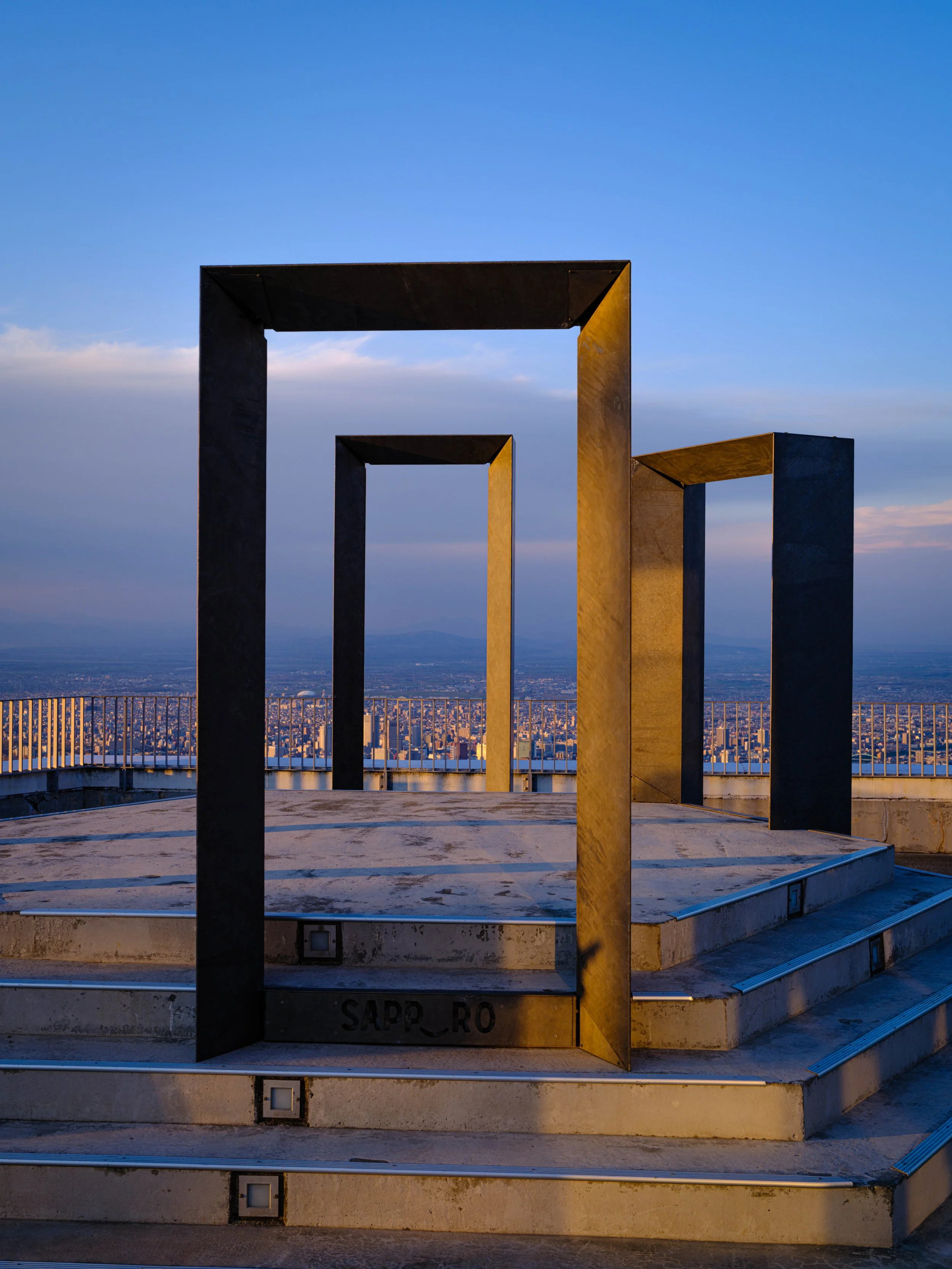 A large metal art sculpture composed of three rectangular frames on a rooftop with a view of a city skyline under a blue sky.
