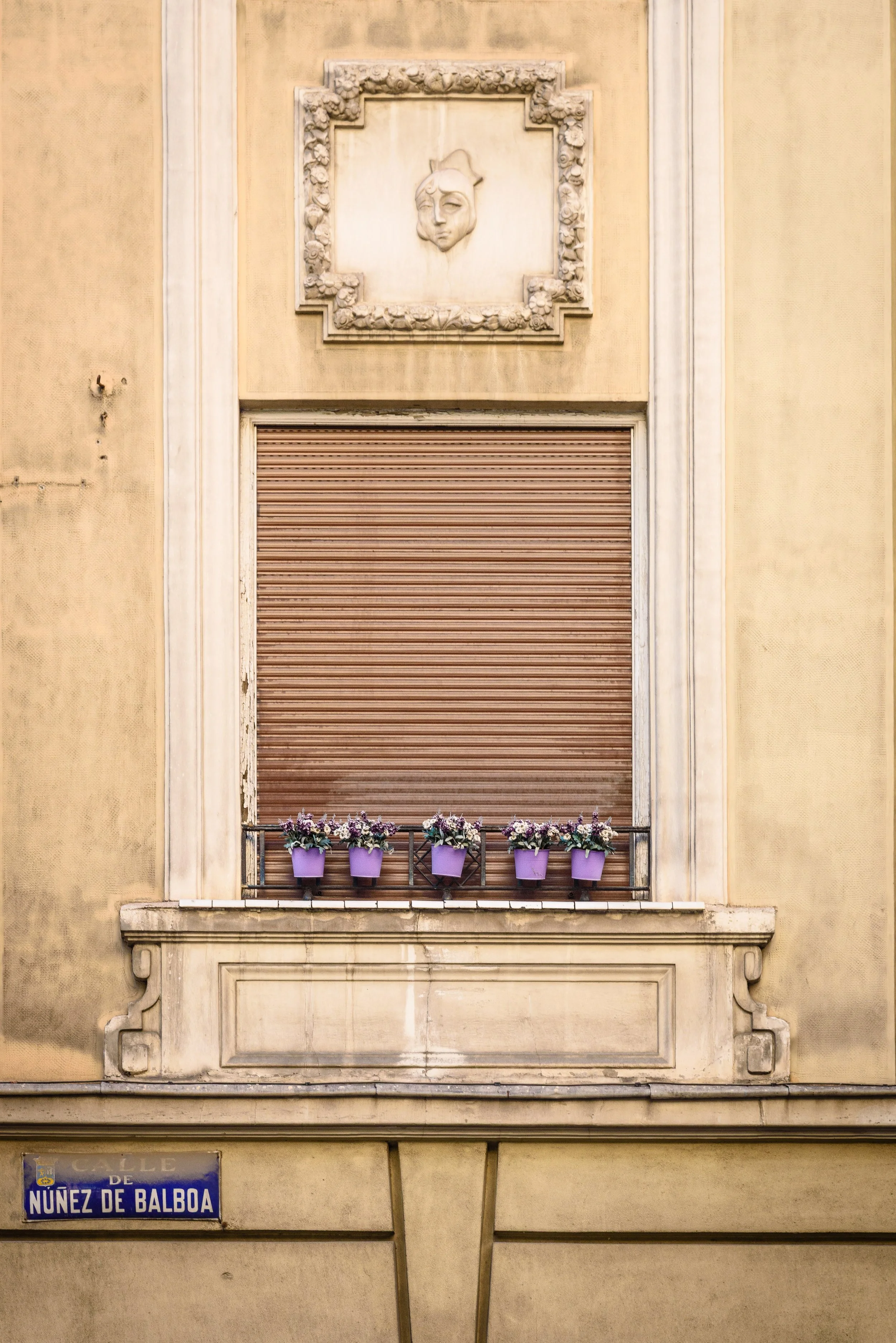A yellow building facade with a closed brown window shutter, adorned with five purple flower pots filled with flowers, and a decorative framed face sculpture above the window. There is a street sign at the bottom left that reads 'Calle de Nunez de Ba