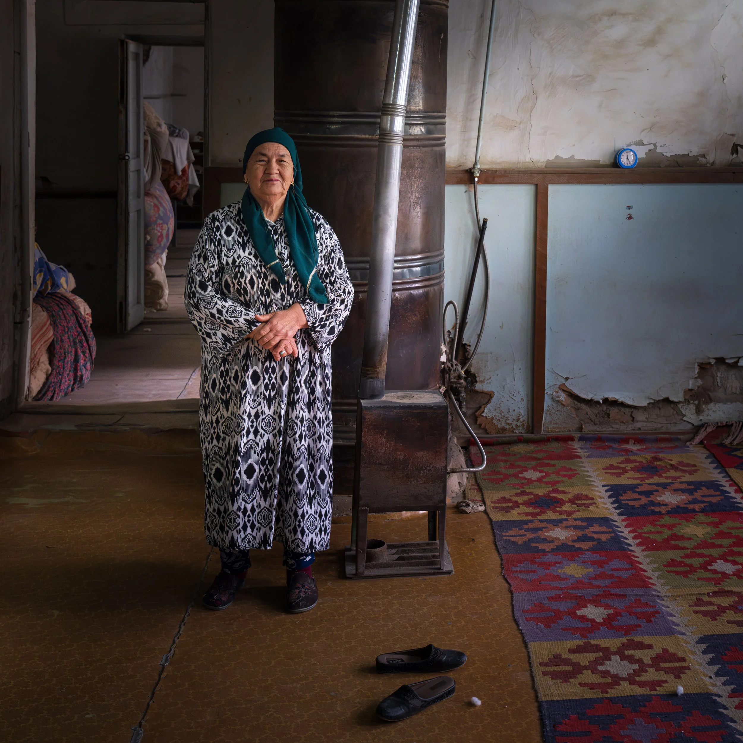An elderly woman standing inside a rustic room with worn walls, standing next to a wood stove, wearing a patterned long dress and teal headscarf, with slippers on the floor nearby.
