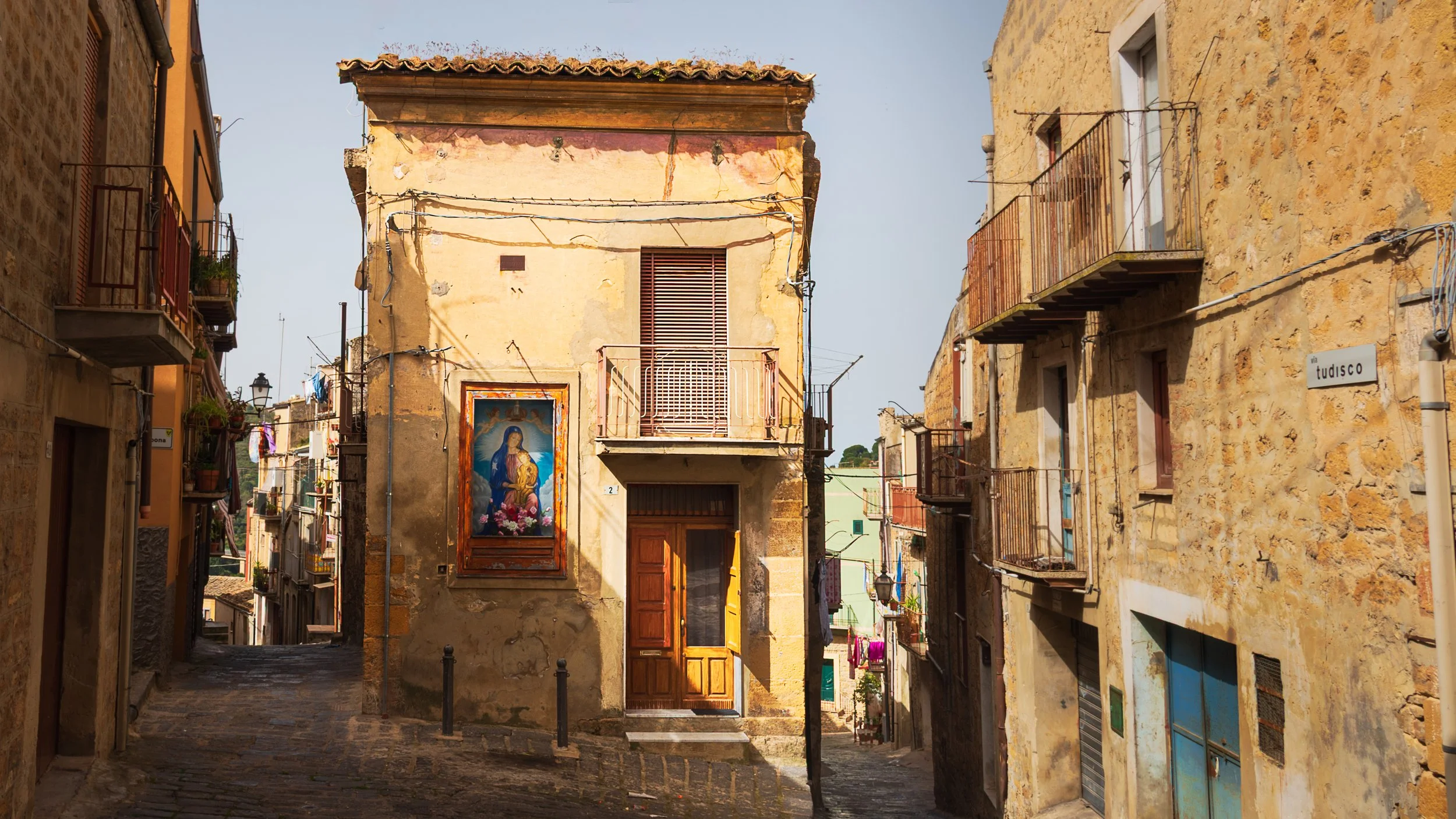 Narrow cobblestone street in a historic European neighborhood with aging buildings, balconies, colorful plants, and a religious painting on the wall.