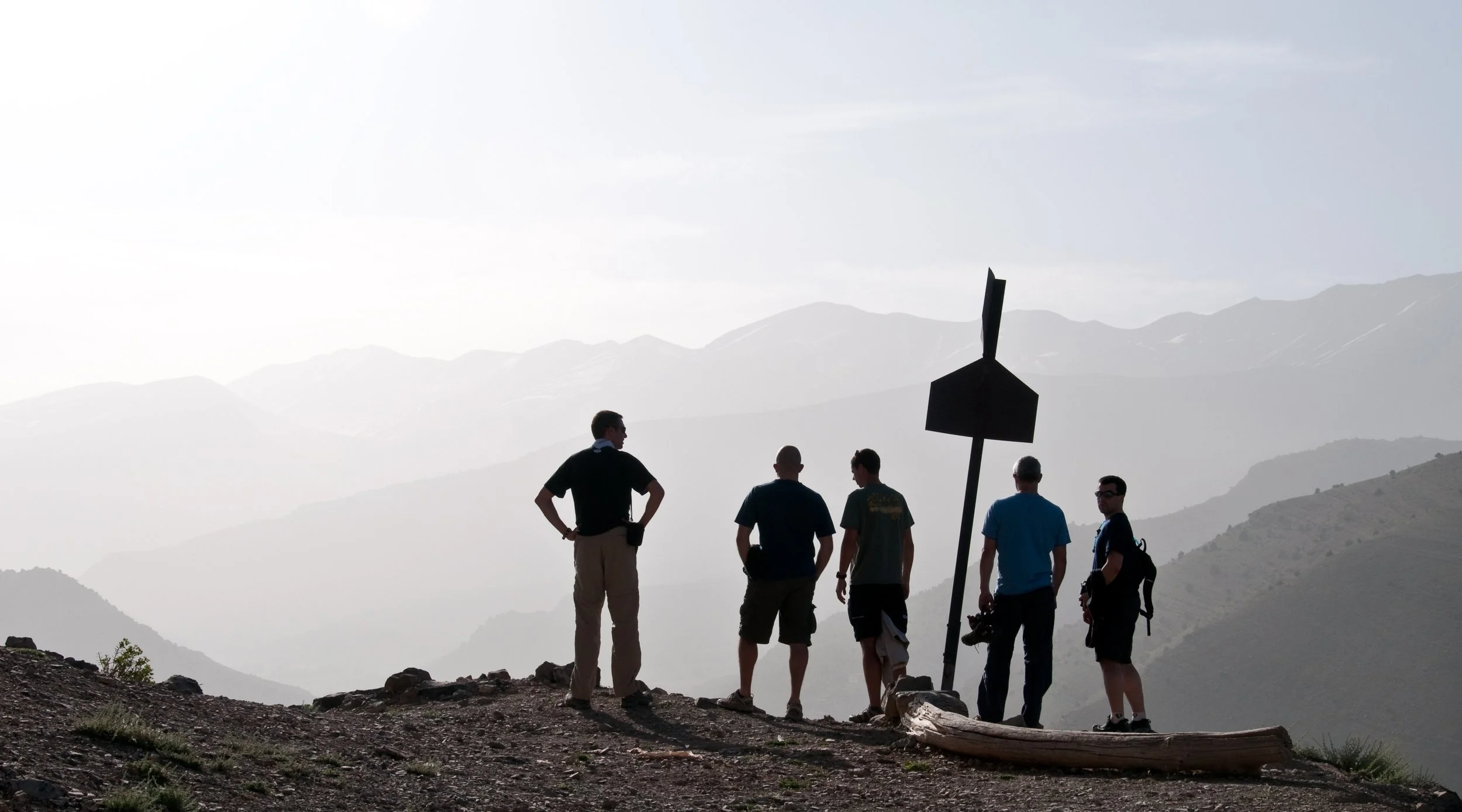Six people standing on a mountain overlook, silhouetted against a hazy mountain range background, with some holding cameras.