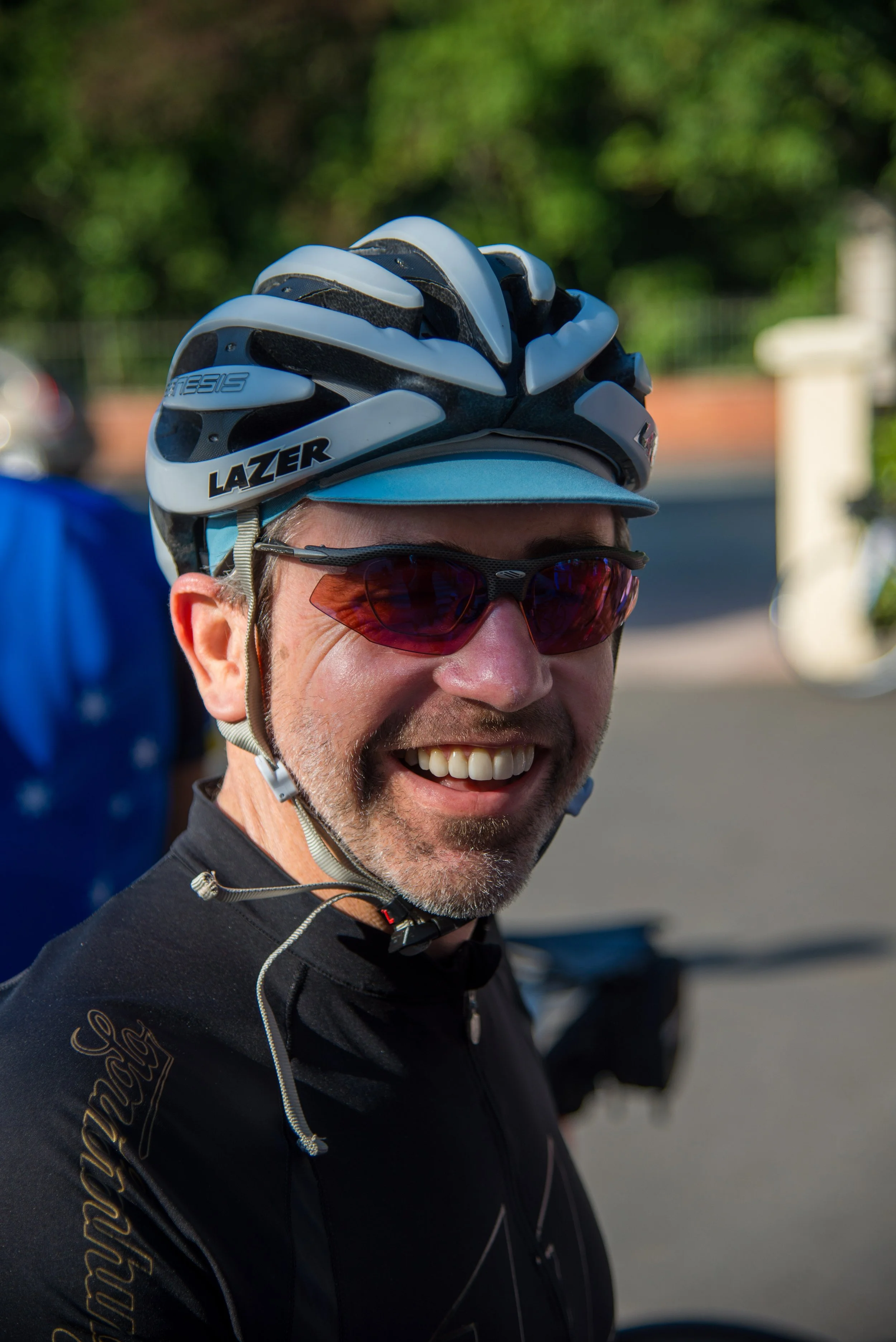 A smiling man wearing a bicycle helmet, sunglasses, and a black cycling jersey outdoors.
