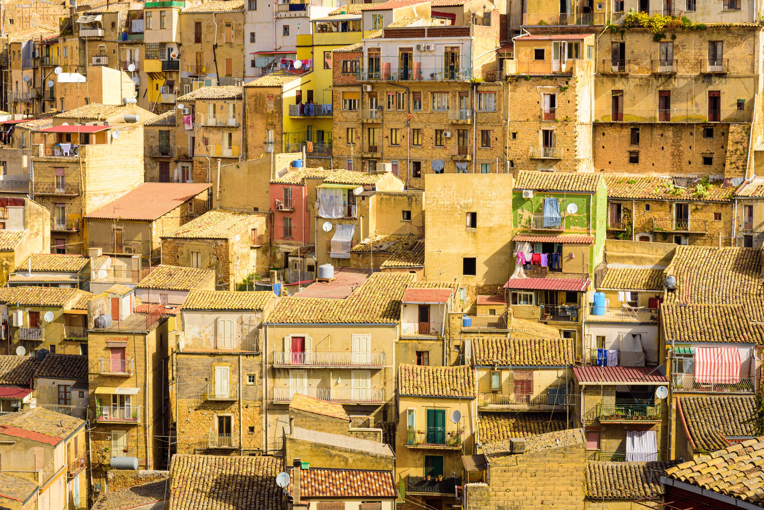 A densely packed hillside of colorful apartment buildings with tiled roofs, balconies, and laundry hanging outside, in a Mediterranean town.
