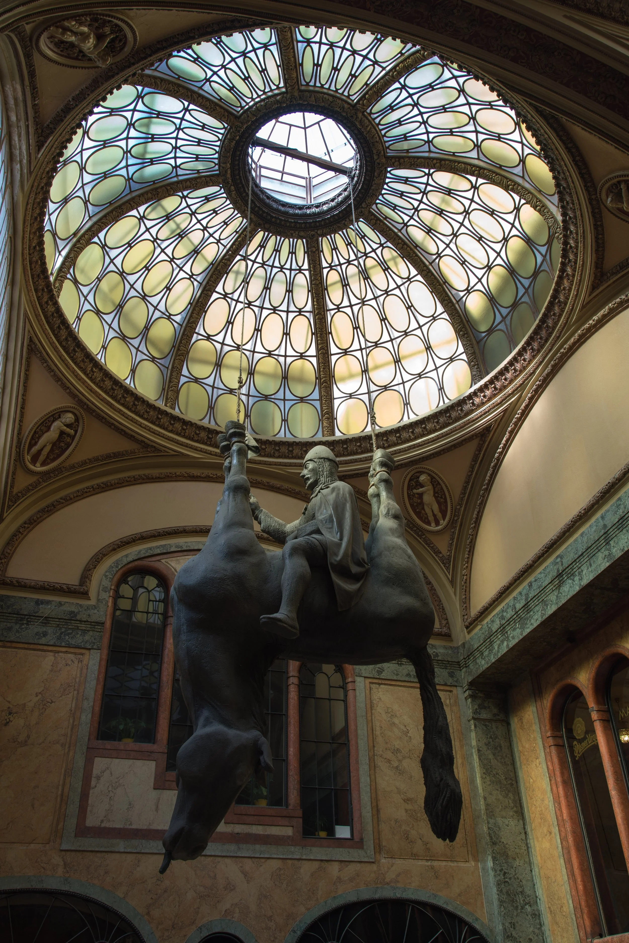 A museum interior featuring a decorative stained glass dome ceiling and a sculpture of a man riding a large, long animal.