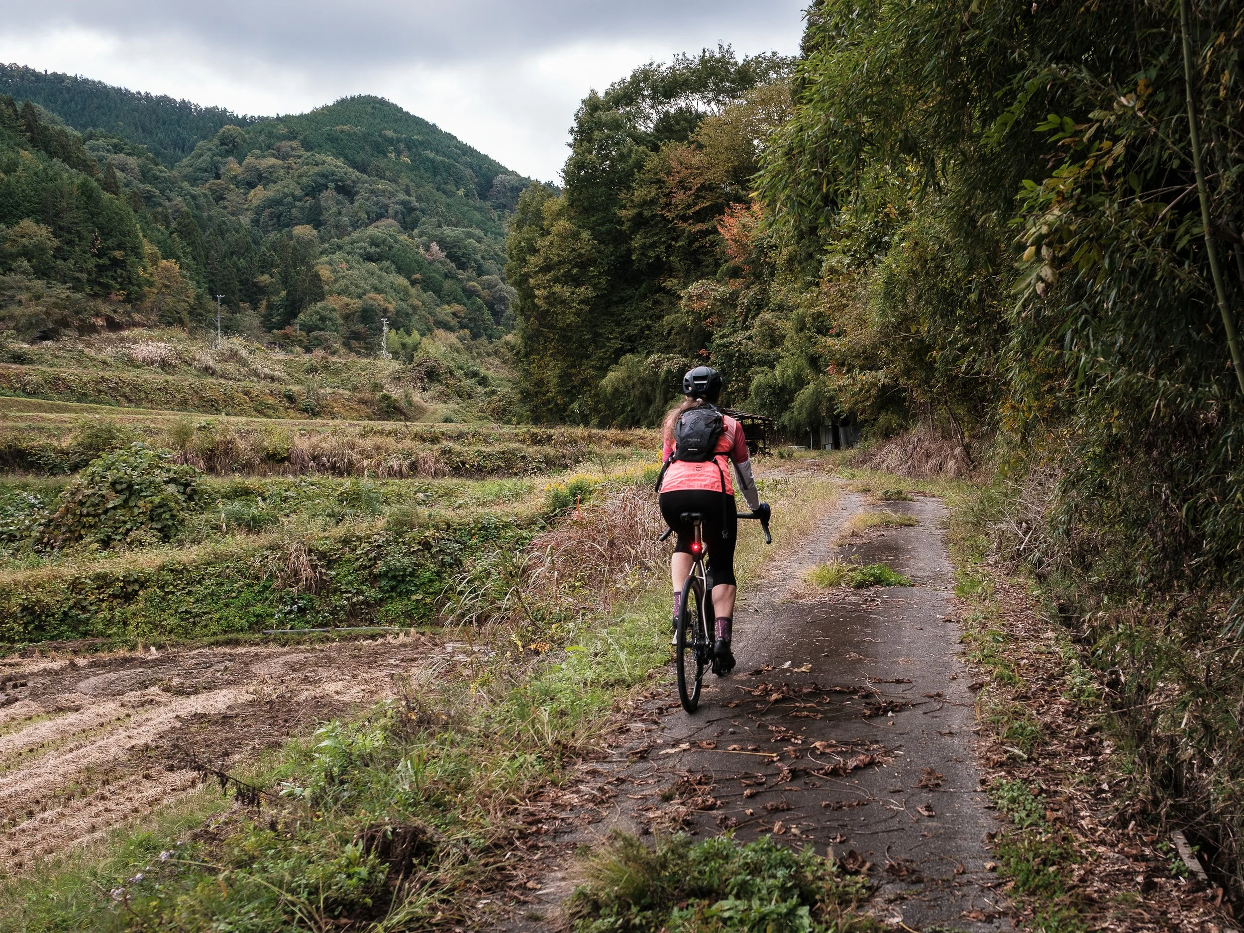 A woman riding a bicycle down a rural dirt and paved trail surrounded by greenery, trees, and hills in the background.