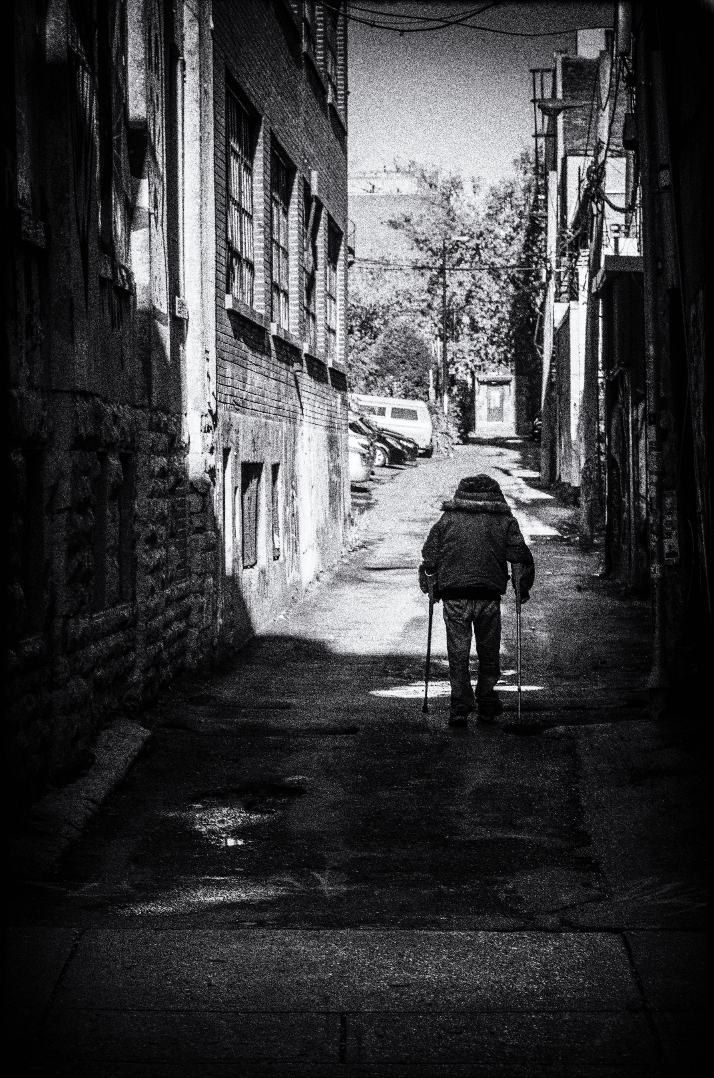 A black and white photo of an elderly person walking with crutches down a narrow alleyway, with buildings on either side and cars parked in the background, heading toward a brighter area with trees.
