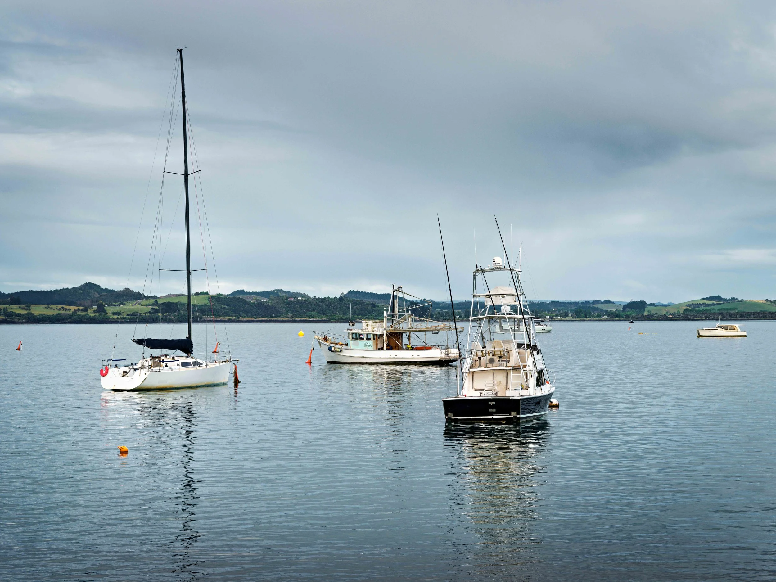 Boats anchored on a calm body of water with hills in the background under a cloudy sky.