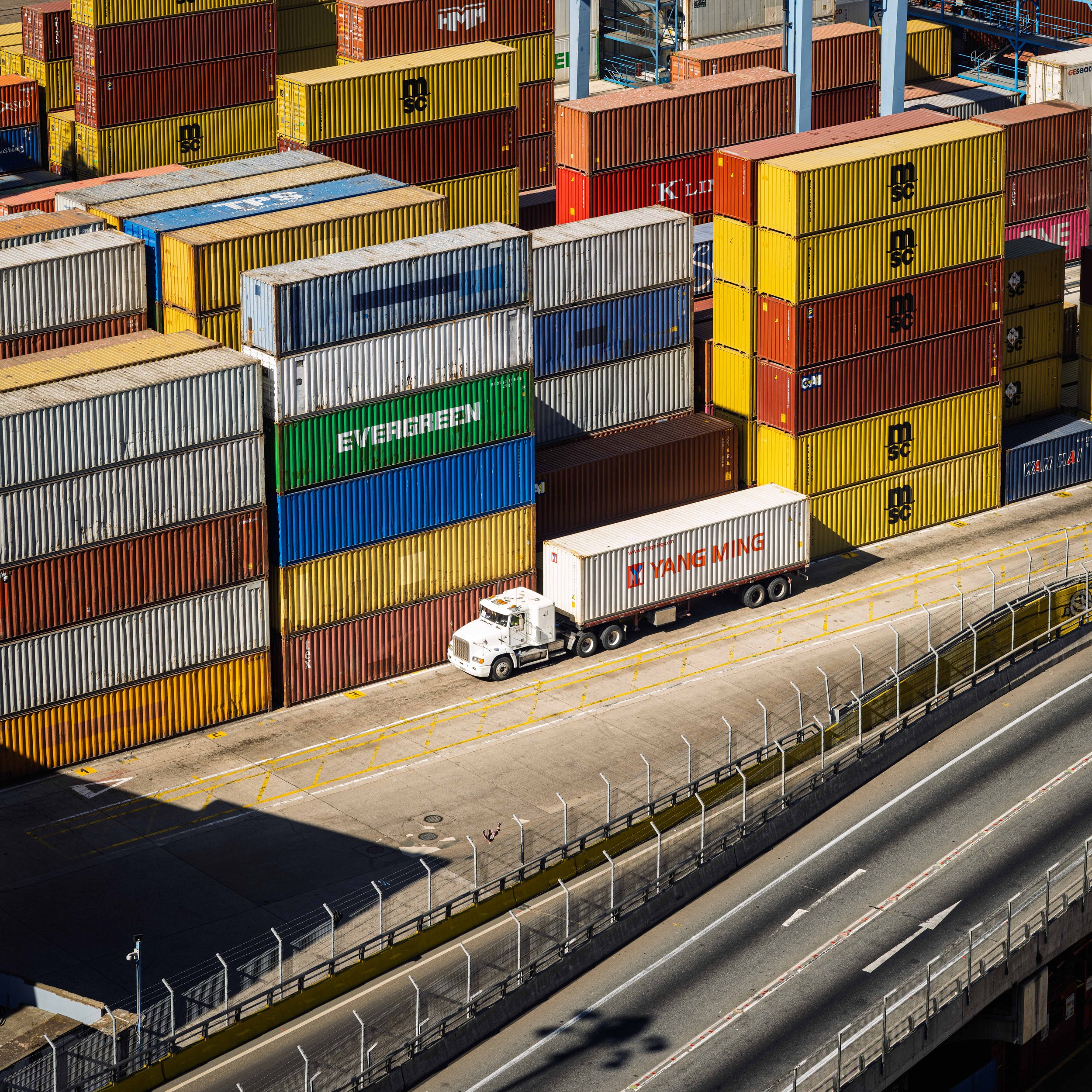 A cargo shipping yard with stacked colorful shipping containers and a white semi-truck on the road beside them.
