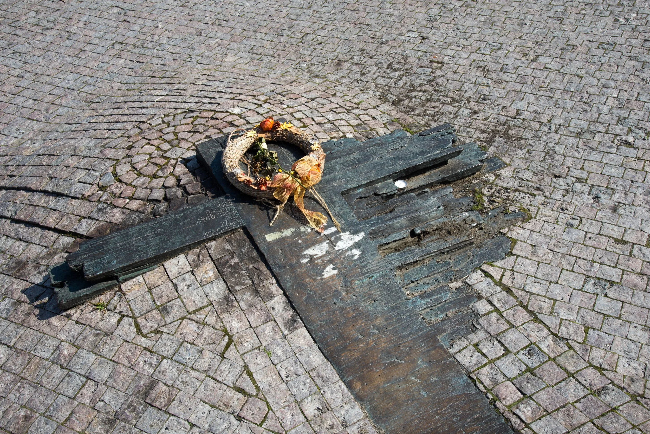 A damaged memorial plaque on a cobblestone ground with a wreath placed on it.