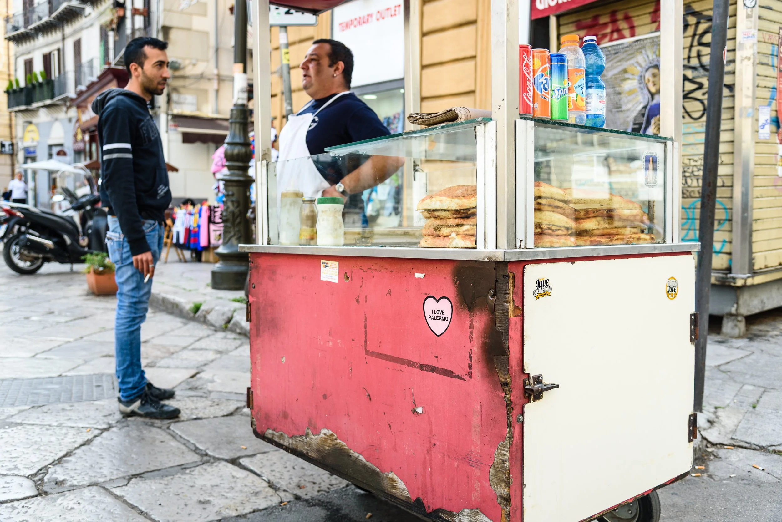 A street vendor cart with stacked sandwiches and beverages, man selling to a young male customer on a city sidewalk.