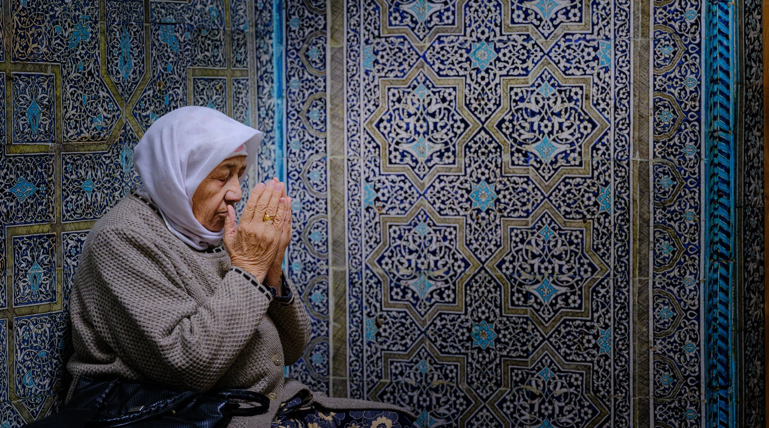 An elderly woman wearing a white headscarf prays with her hands pressed together, sitting against an intricate, colorful tiled wall.