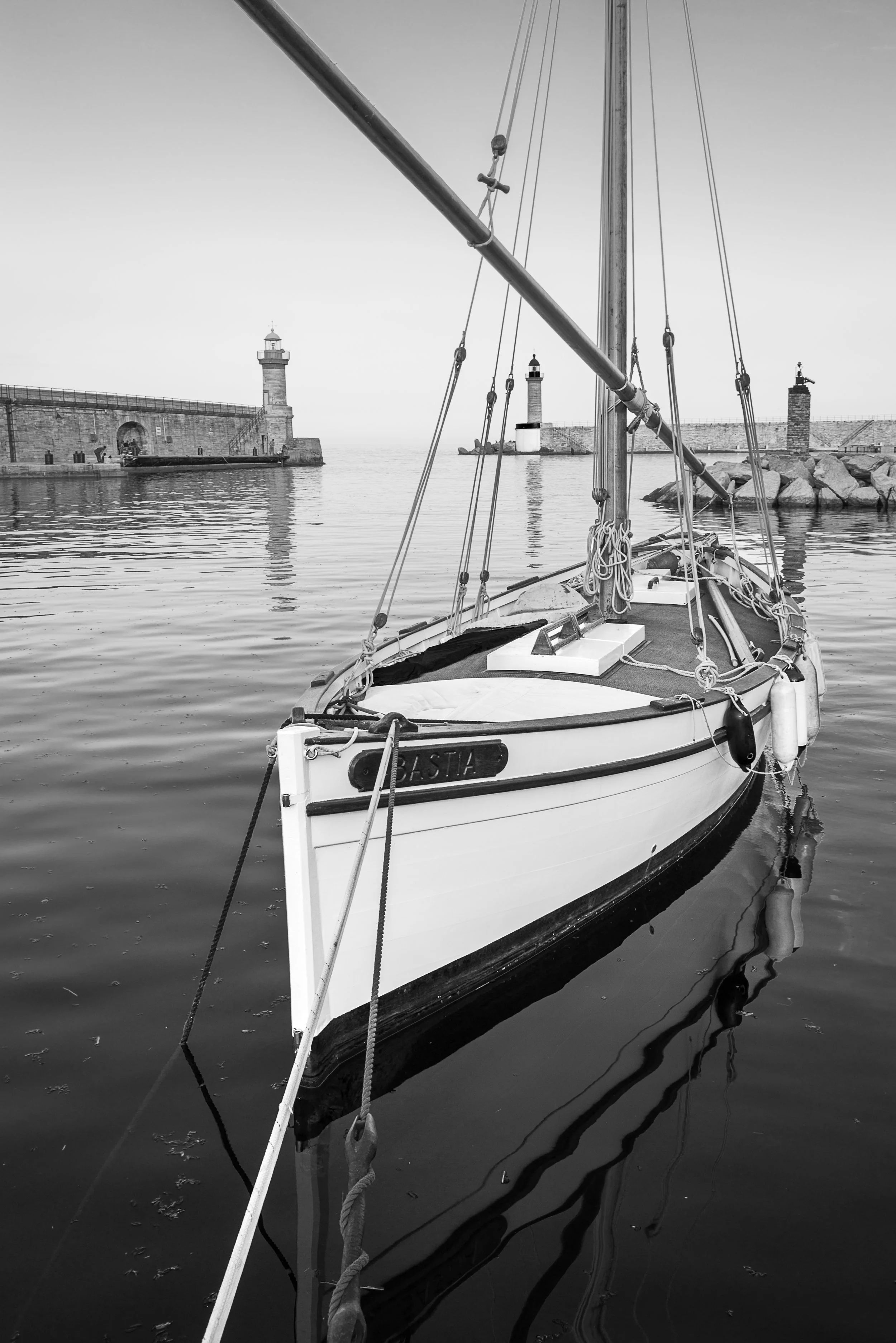 A black and white photo of a sailboat docked in calm water with a lighthouse and stone pier in the background.