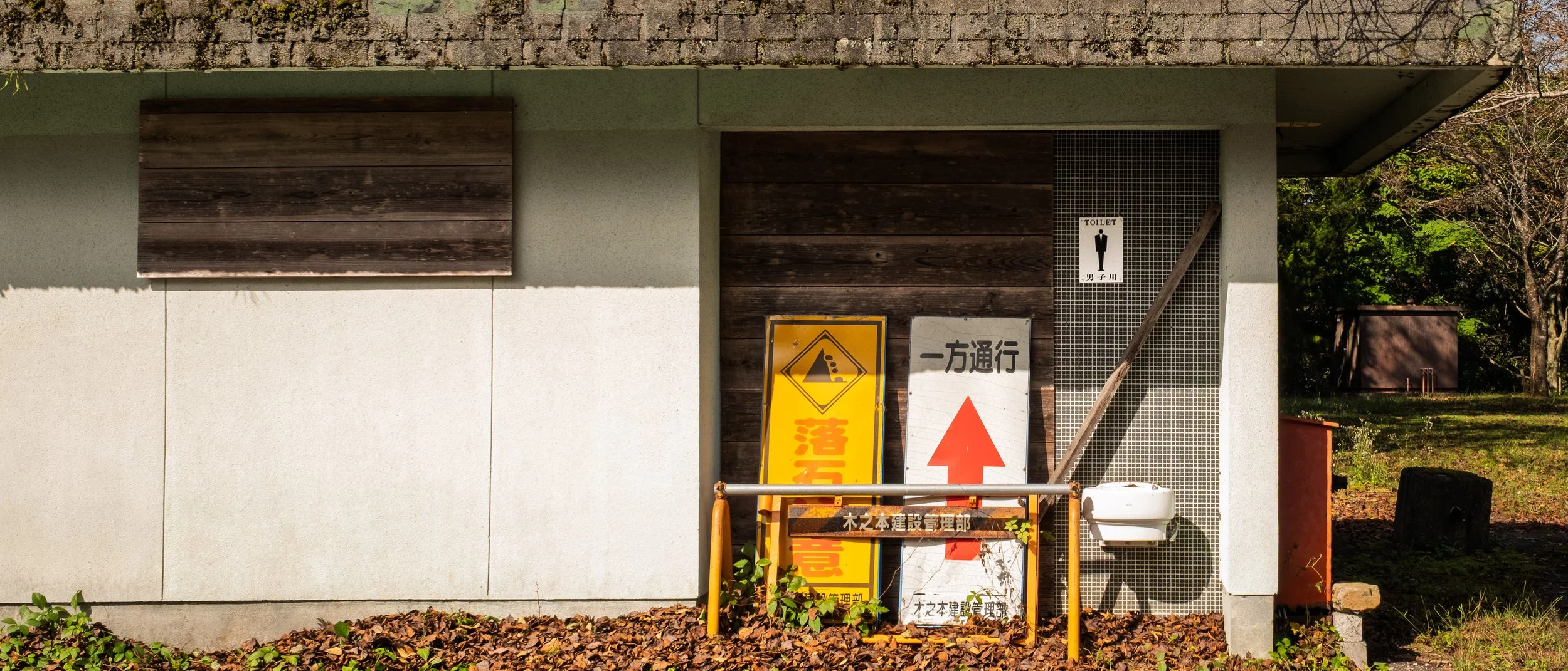 Exterior view of a building with a restroom sign and construction warning signs, including a yellow warning sign with a cave-in symbol, set against a background of trees and grass.