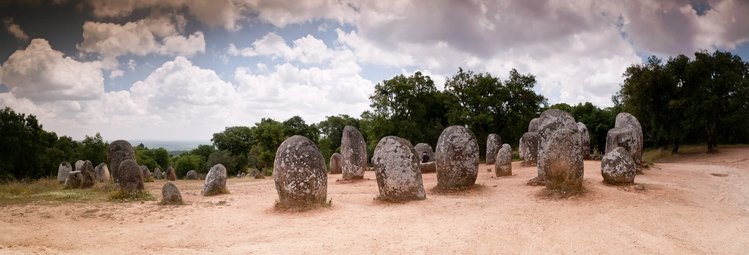 A prehistoric stone circle with large, weathered standing stones arranged on a sandy terrain under a partly cloudy sky, surrounded by green trees.