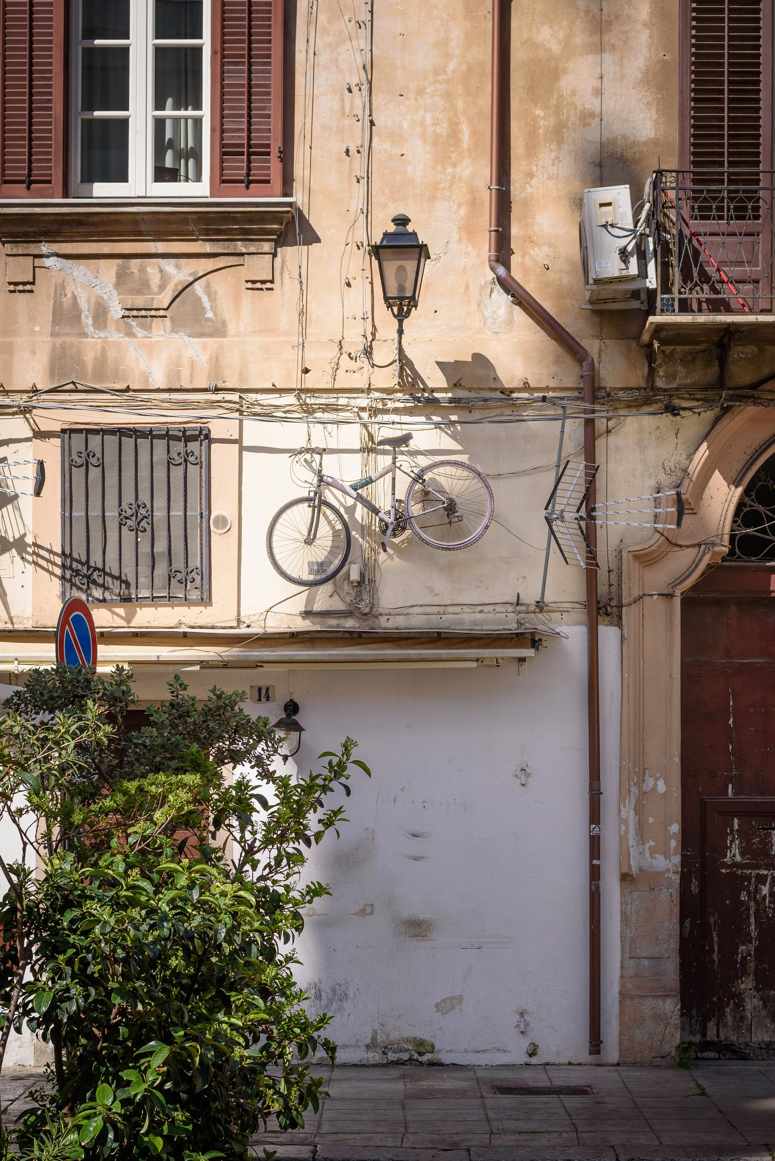 Old building wall with a mounted bicycle, a street lamp, wires, windows with shutters, an air conditioning unit, a small balcony, and lush greenery.