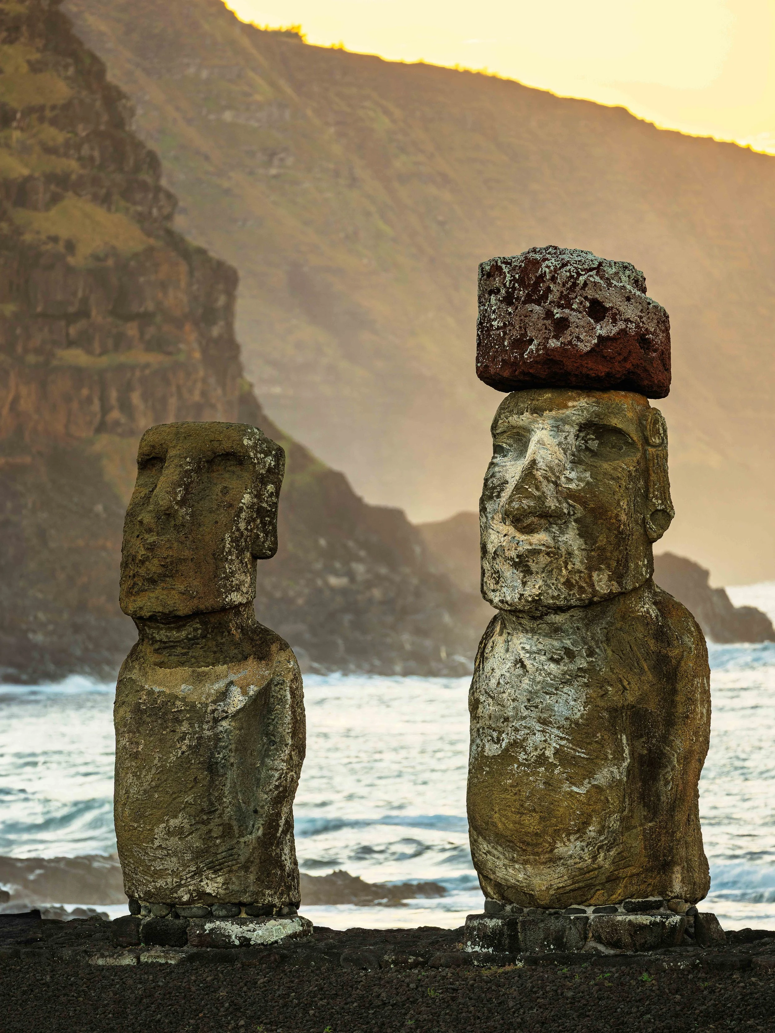 Two Moai statues on a beach with cliffs in the background and a sunset sky.