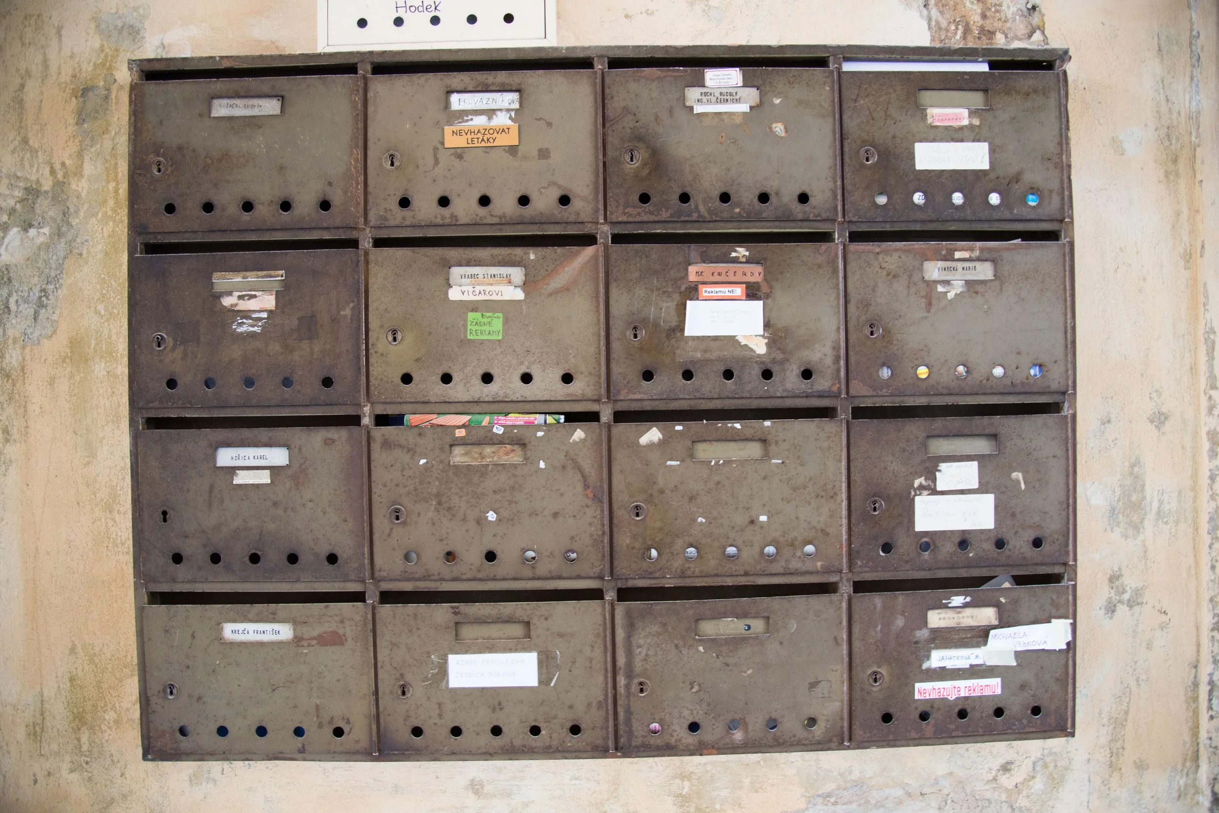 A weathered metal mailboxes with various labels and notices, mounted on an aged, peeling wall.