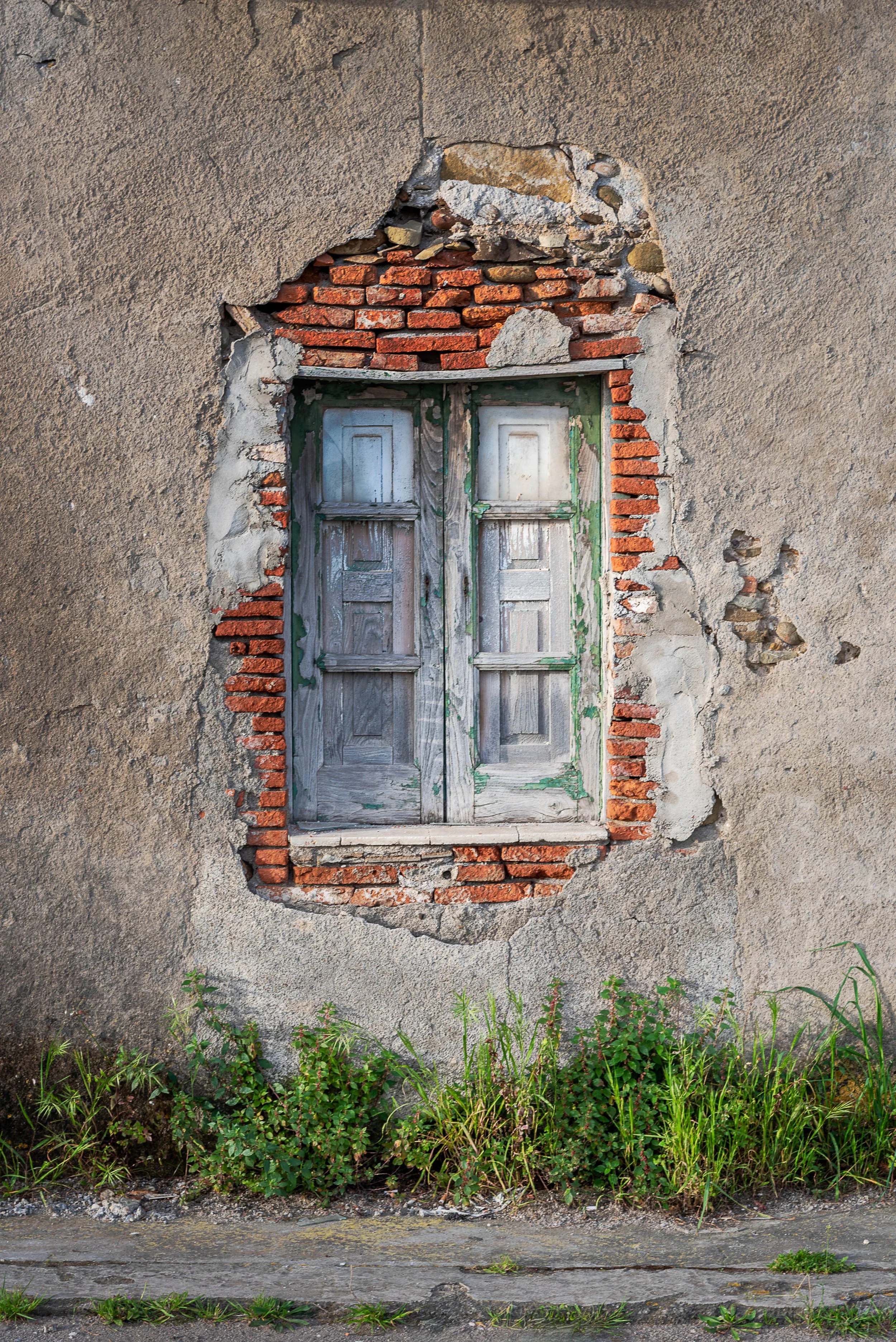 Old, weathered window with wooden shutters in a damaged brick and concrete wall, with grass in front.