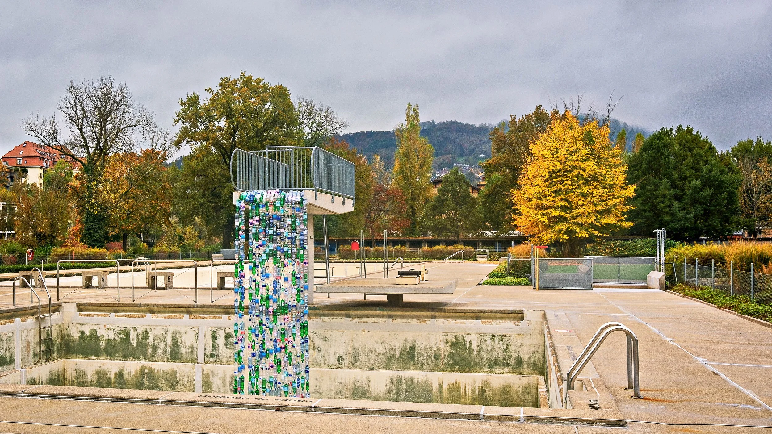 Empty outdoor swimming pool with a waterfall made of plastic bottles, surrounded by a fenced deck with trees and houses in the background on a cloudy day.