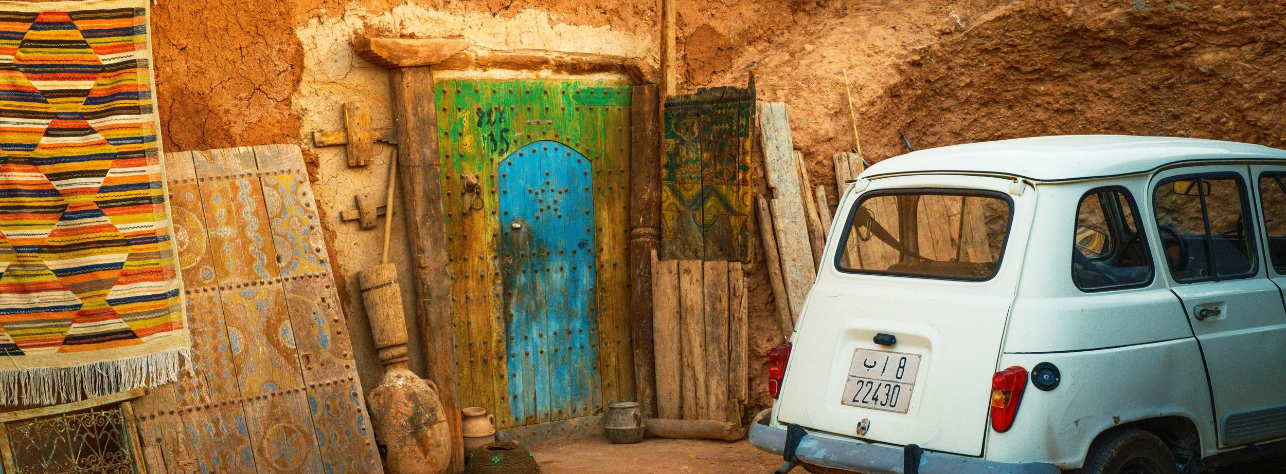 A rustic scene with a vintage white car parked next to a wall decorated with traditional textiles, wooden doors, and artifacts against a rough, adobe wall.