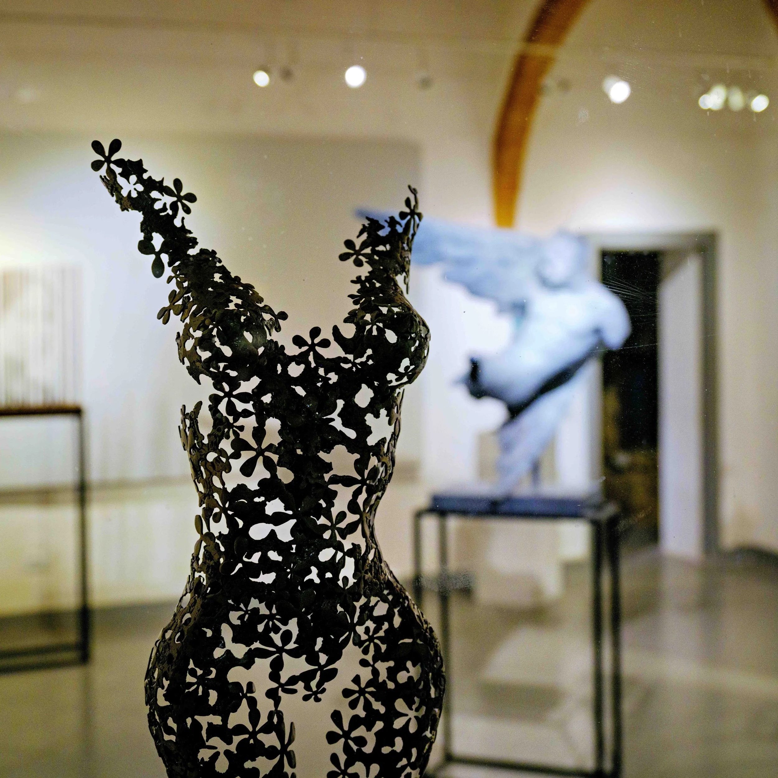 Metal sculpture of a female torso with floral design in an art gallery, with another sculpture visible in the background.