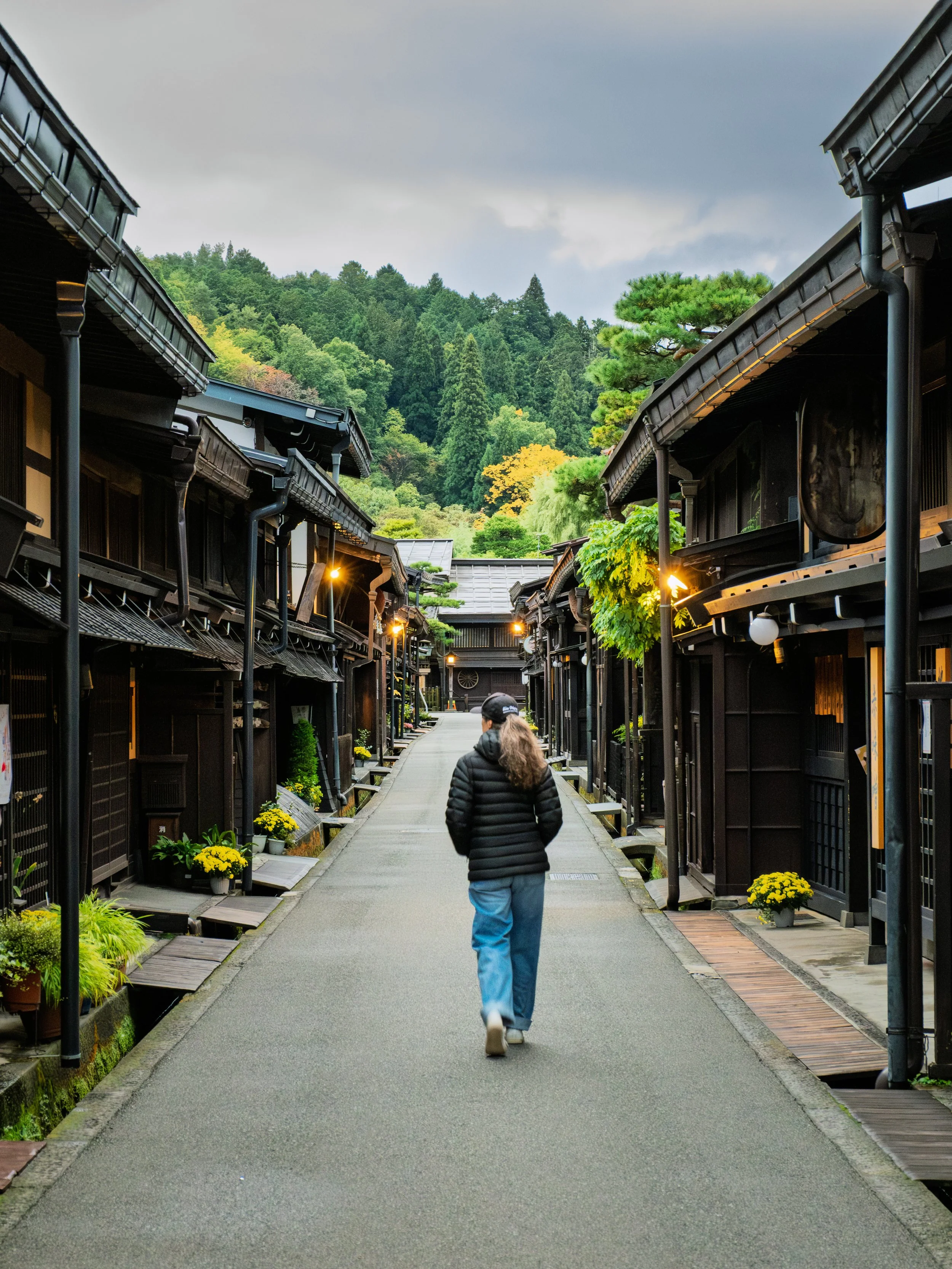 A person walking down a traditional Japanese street lined with dark wooden buildings and yellow potted flowers, with a forested hillside in the background.