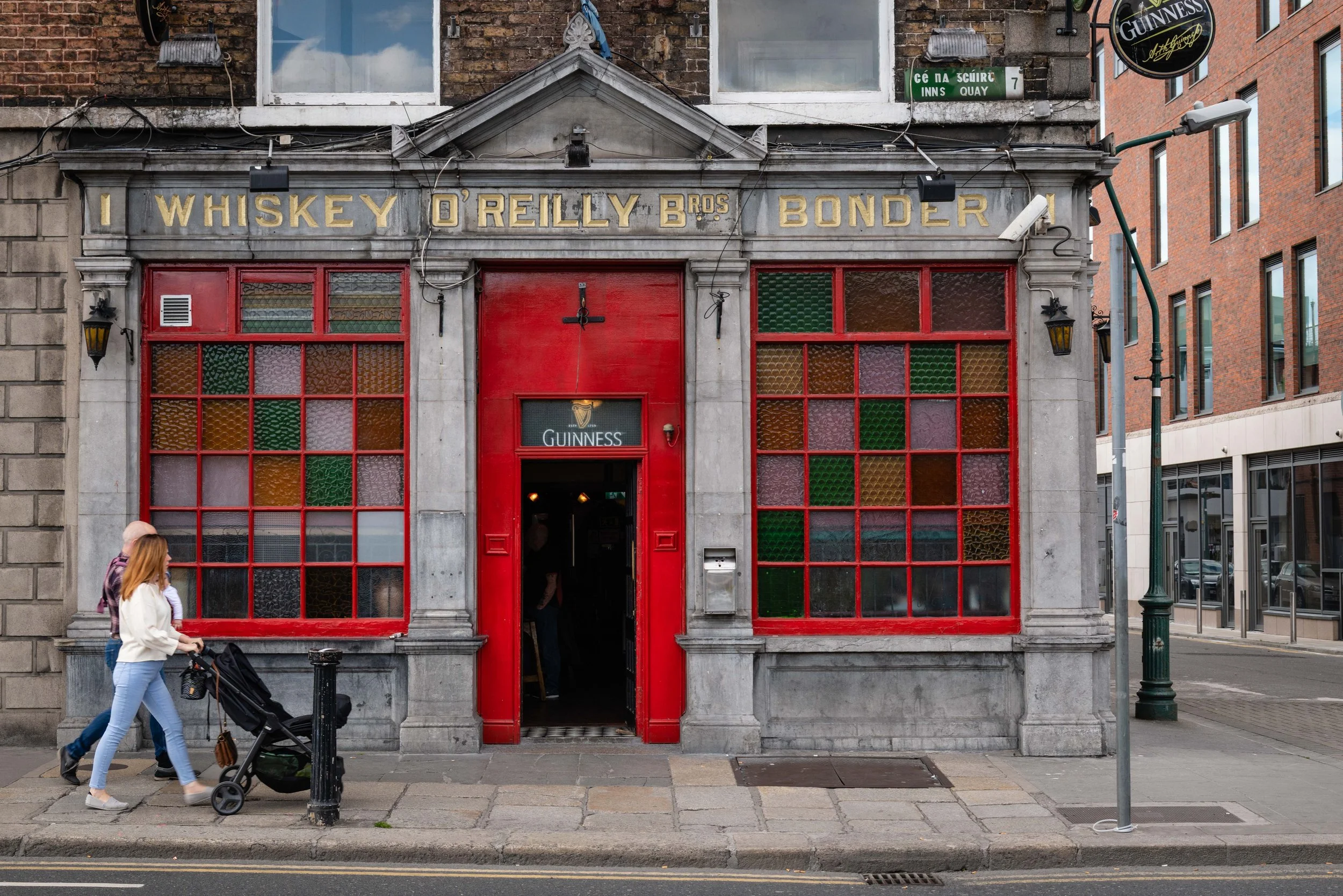 Front view of a pub with red doors and stained glass windows, called Whiskey O'Reilly Bros Bonder, on a city street with a woman pushing a stroller and a man walking nearby.