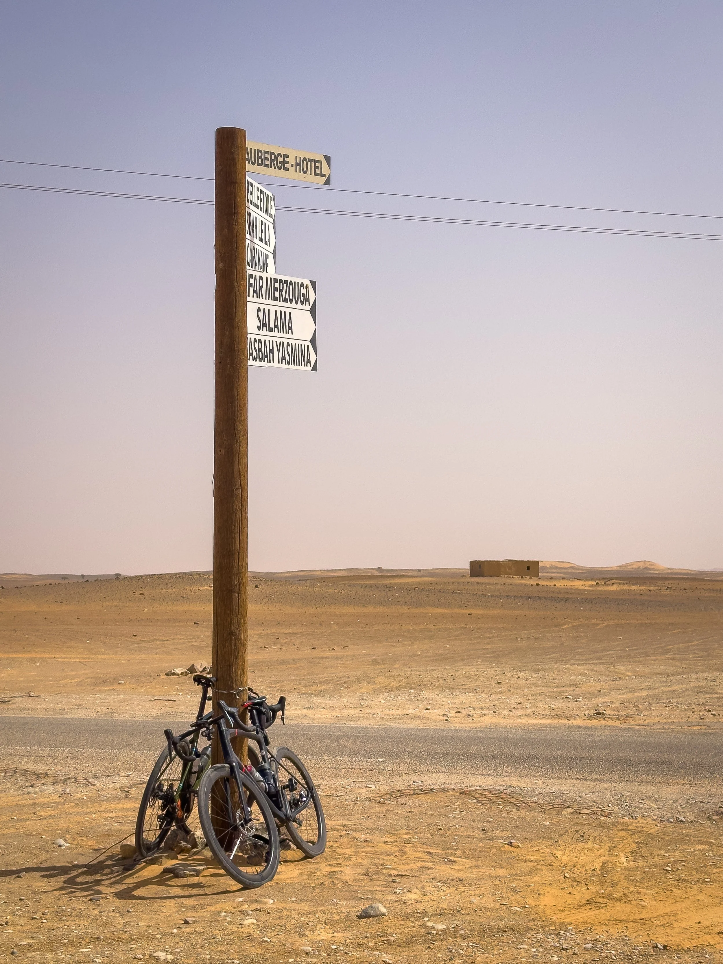 Two bicycles leaning against a wooden utility pole in a desert landscape with a signpost with multiple directional signs, including one labeled 'Auberge Hotel'.