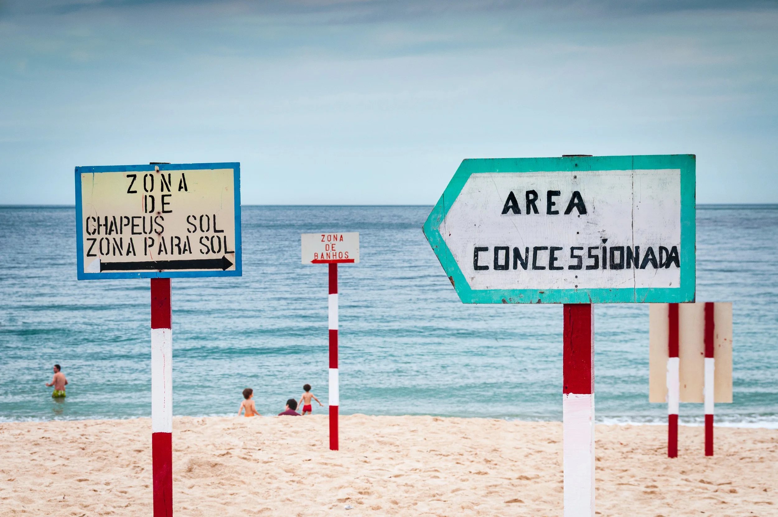 Beach with multiple signboards in Portuguese and Spanish, people swimming and relaxing in the ocean, sandy shore, and cloudy sky.