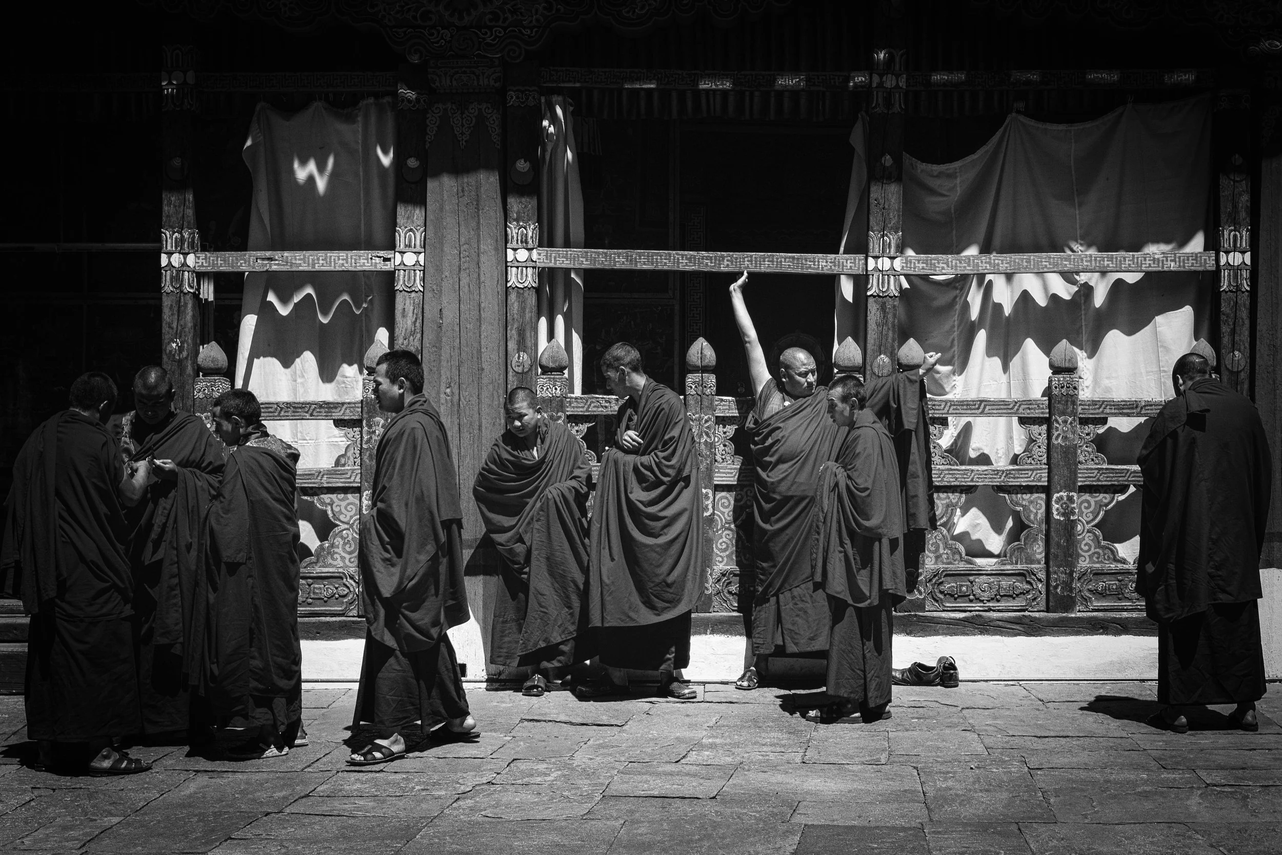 Monks in traditional robes standing and engaging in prayer outside a temple with ornate woodwork and draped fabric in black and white.