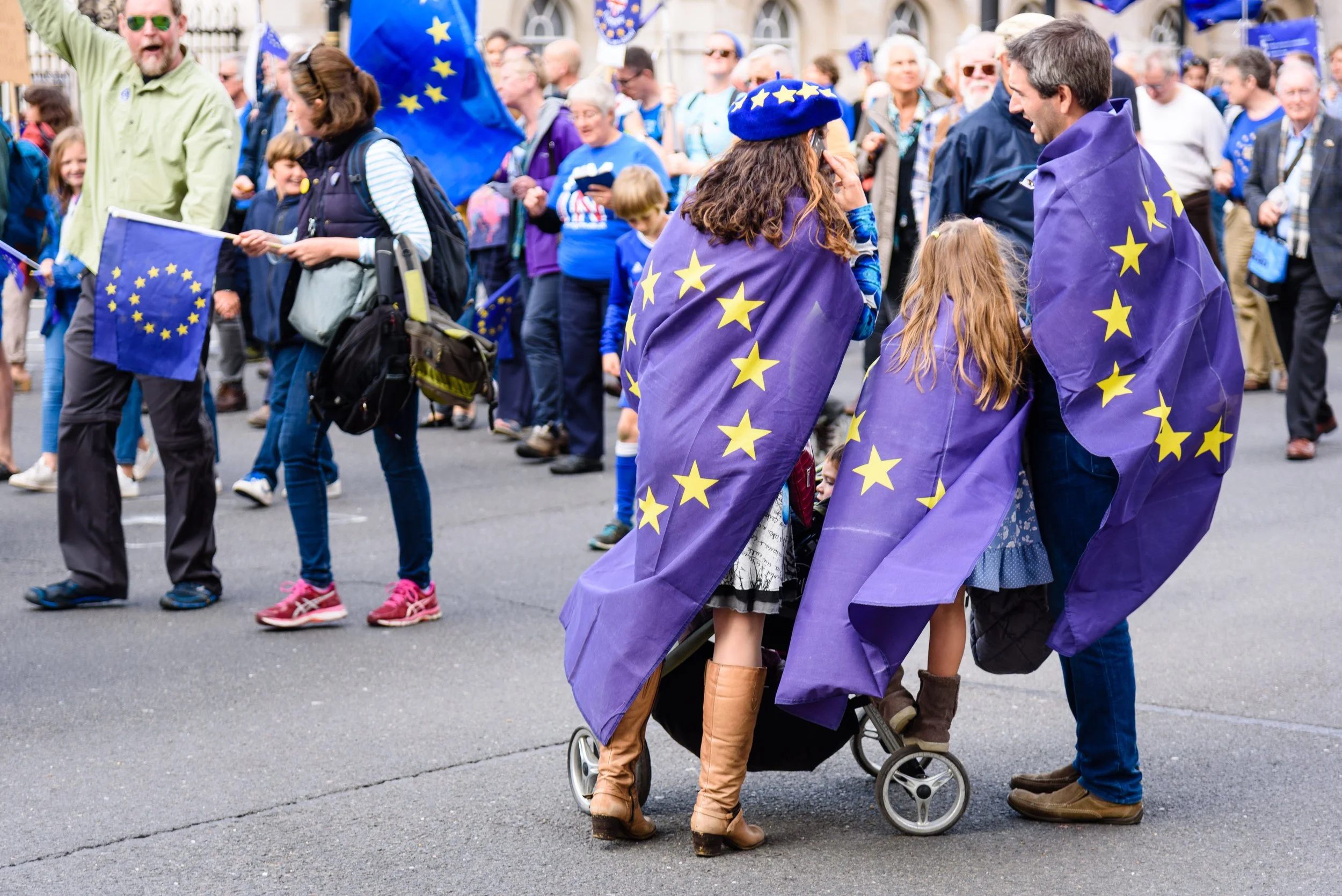 Crowd of people participating in a demonstration or gathering, many wearing or holding European Union flags, with some people wearing EU-themed capes and hats.